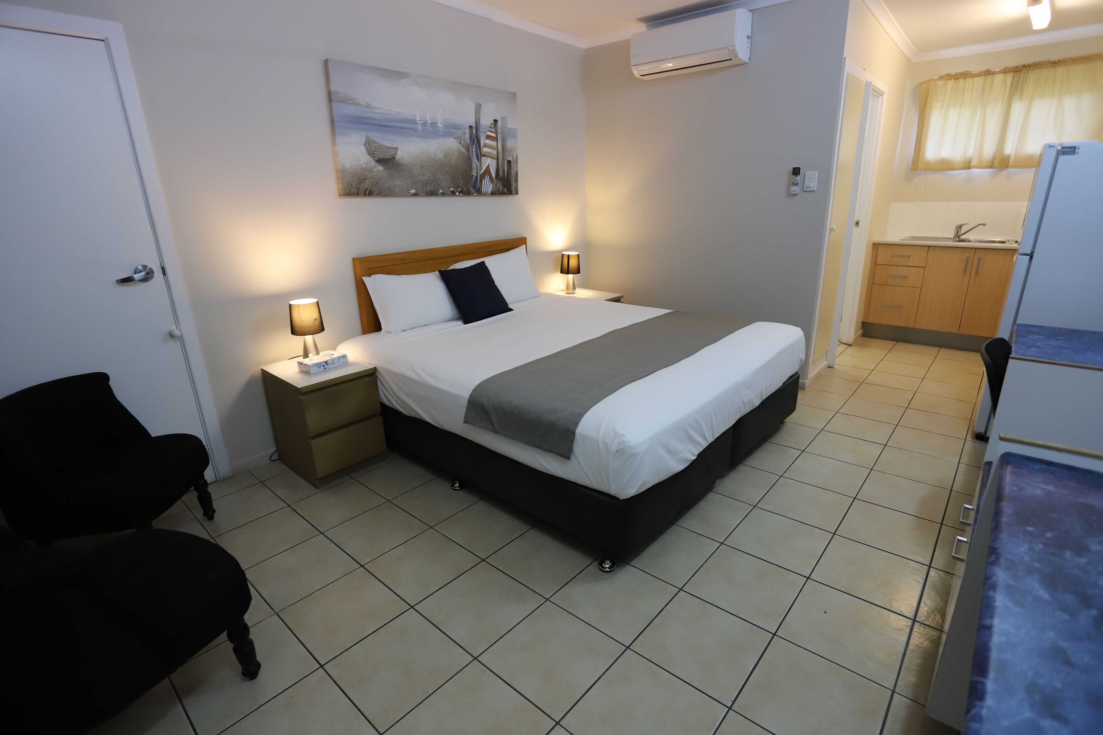 A bedroom with a bed, bedside tables, a chair, a painting, and a kitchenette.