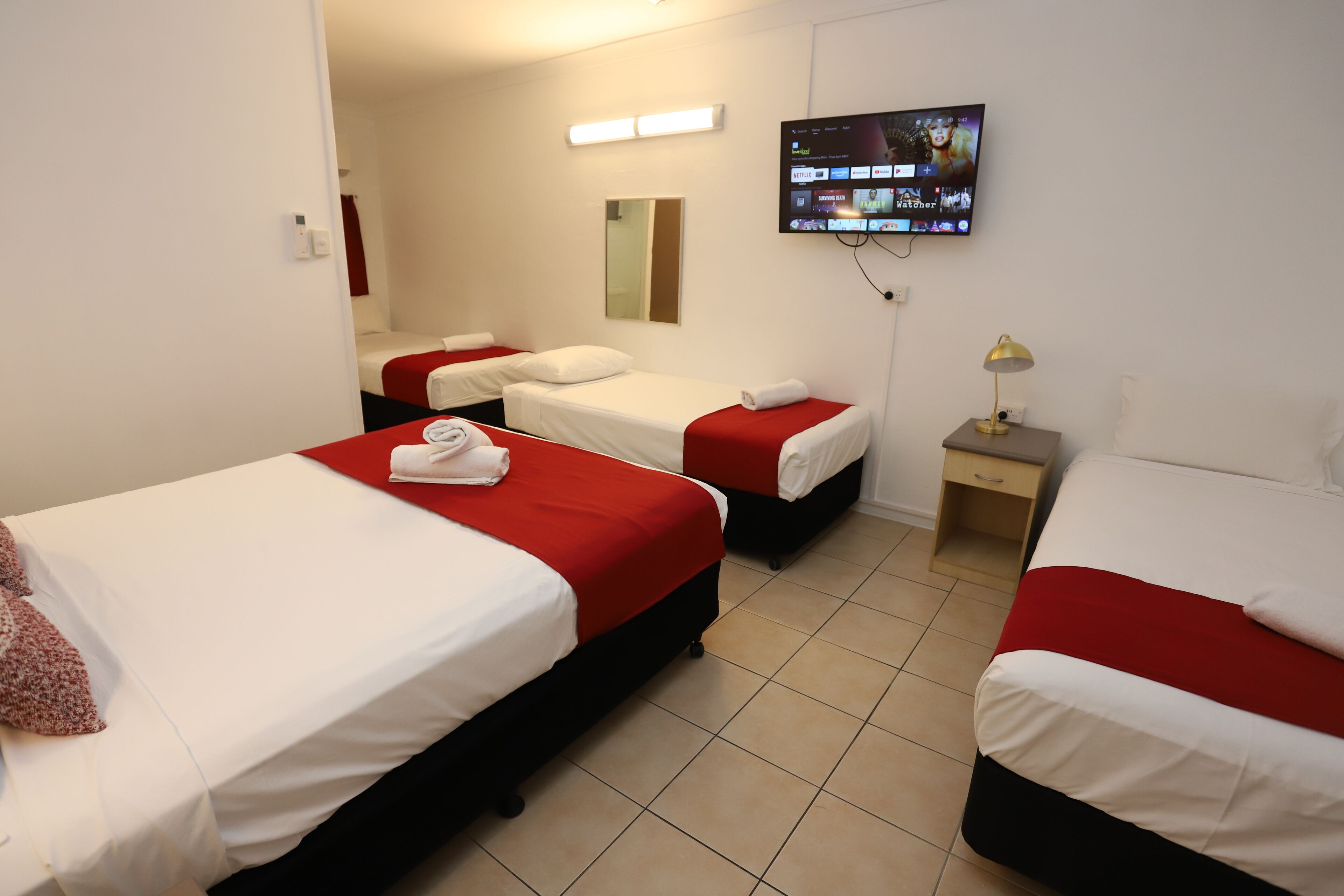 Une chambre d’hôtel avec trois lits, chacun doté de draps rouges et blancs, un téléviseur à écran plat fixé au mur et une petite table de chevet avec une lampe.