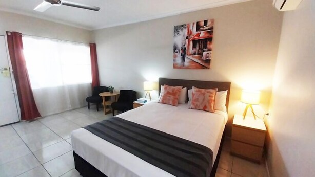 A hotel room with a large bed, two bedside lamps, a ceiling fan, a small table, and a chair. - Tropic Coast Motel (West Mackay)