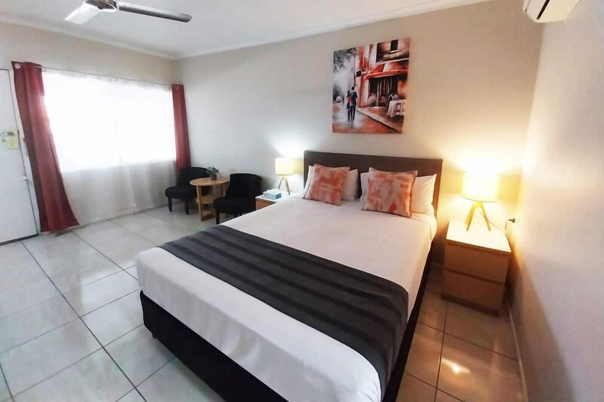 Une chambre d’hôtel avec un grand lit, deux lampes de chevet, un ventilateur de plafond, une petite table et une chaise.