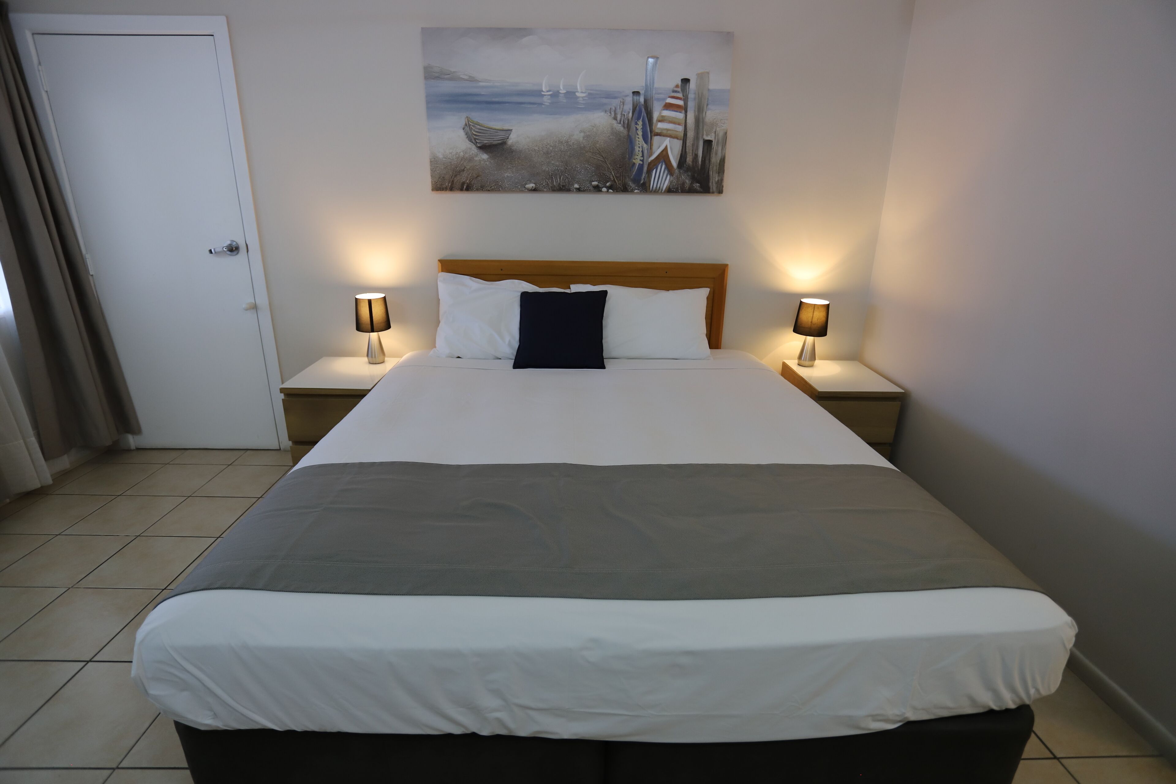 Una habitación de hotel con una cama grande, dos mesitas de noche con lámparas, un cuadro en la pared y una puerta con cortinas.
