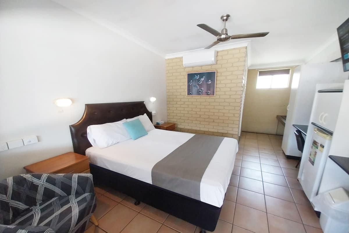 Une chambre d’hôtel comprenant un lit, une chaise, un bureau, un réfrigérateur, un ventilateur de plafond et un mur orné d’un tableau.