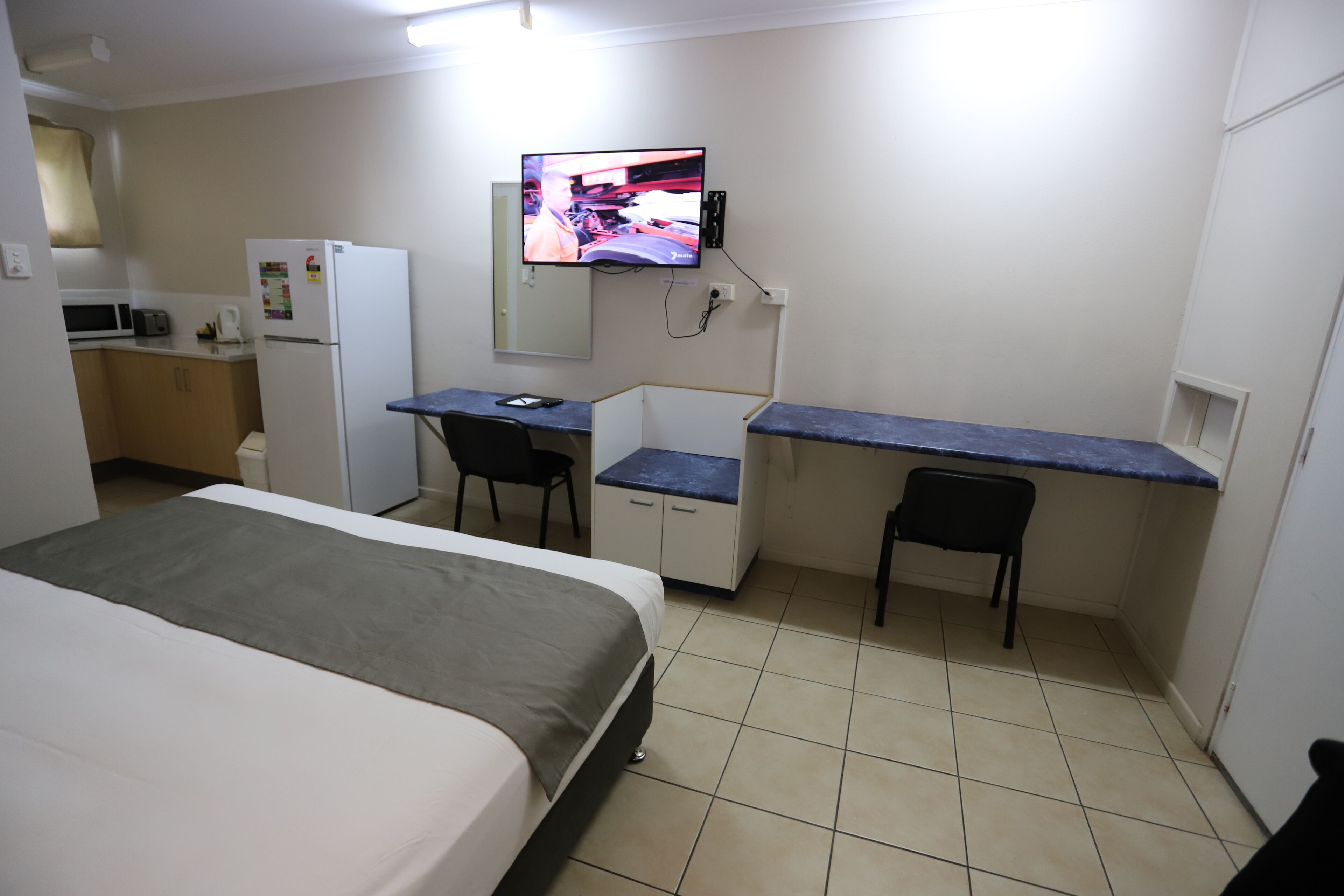 A compact hotel room with a bed, a desk, two chairs, a small kitchenette, and a TV mounted on the wall.