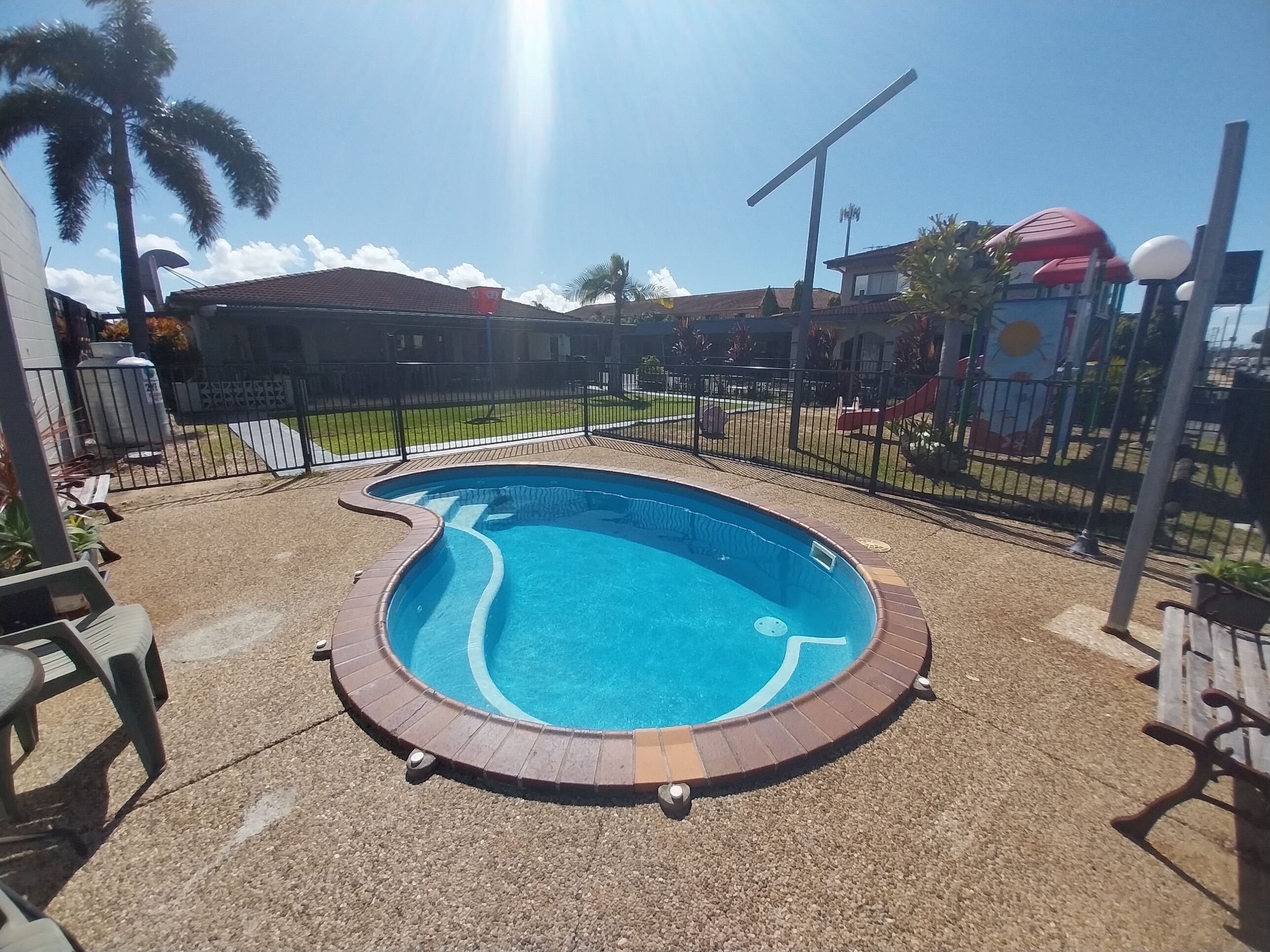 A swimming pool with a brick border, surrounded by a fence, and a playground in the background.