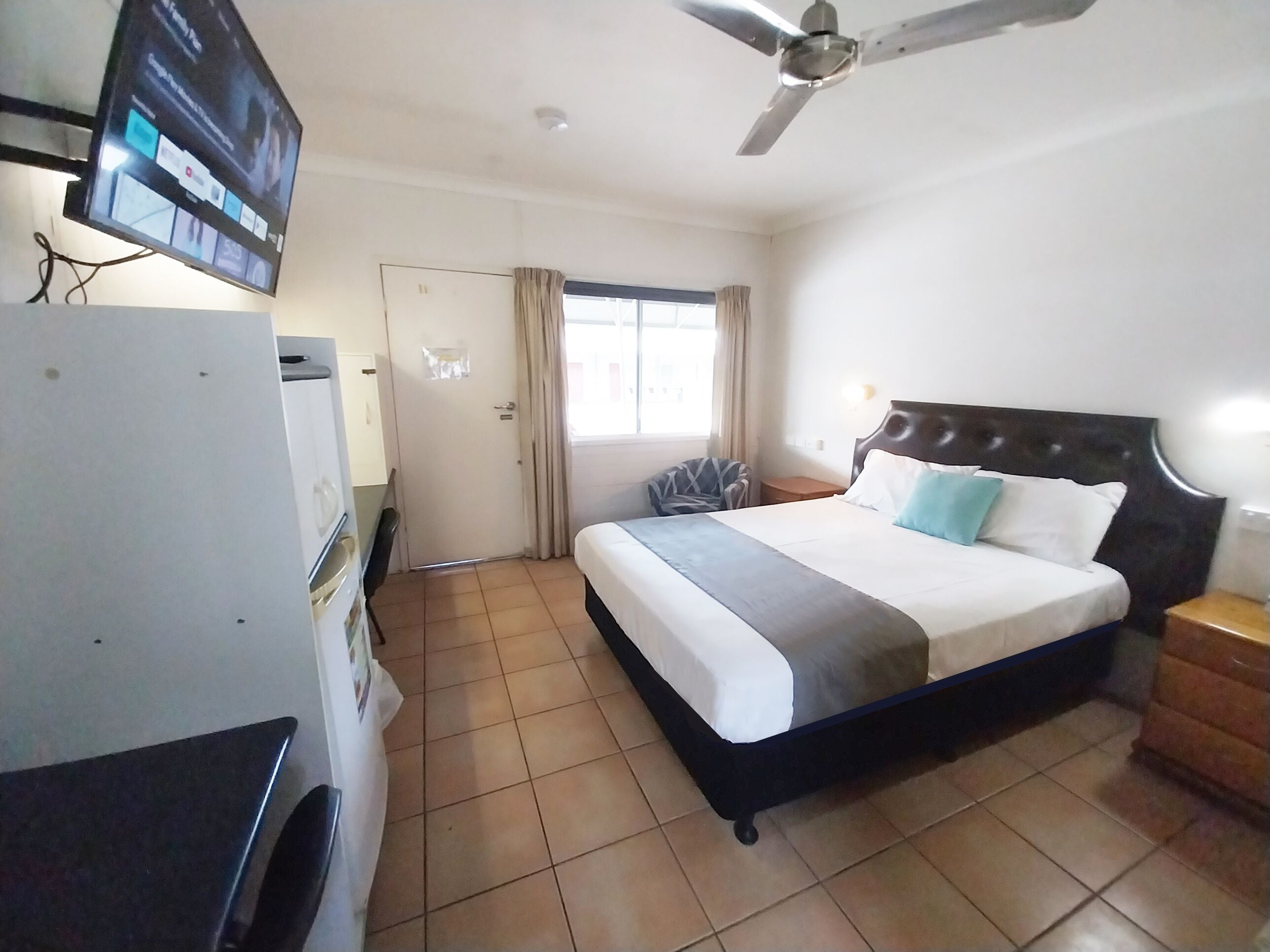 A hotel room with a bed, a TV, a ceiling fan, a window with curtains, and a door.