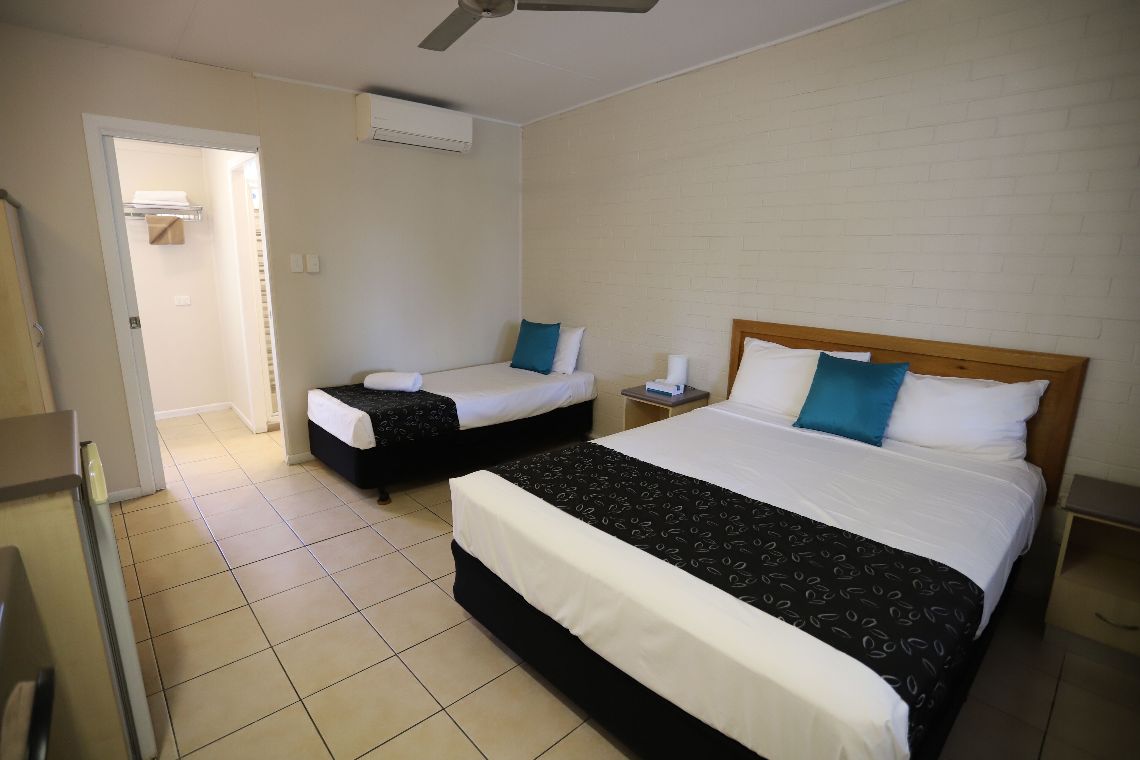 Habitación de hotel con dos camas individuales, una cama grande, un ventilador de techo y un baño visible a través de una puerta abierta.