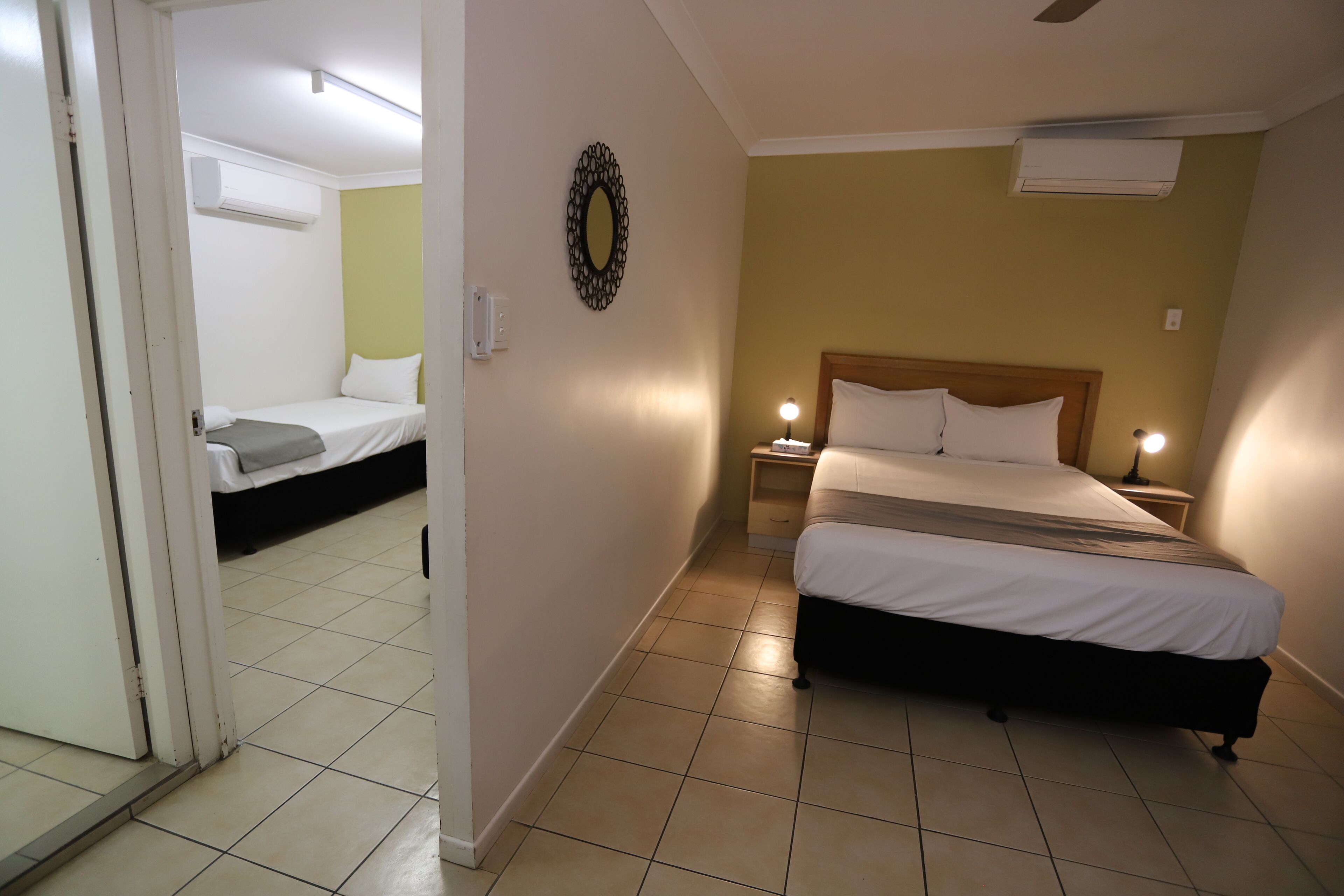 A hotel room with a single bed, bedside tables with lamps, a wall-mounted air conditioner, and a door leading to another room.