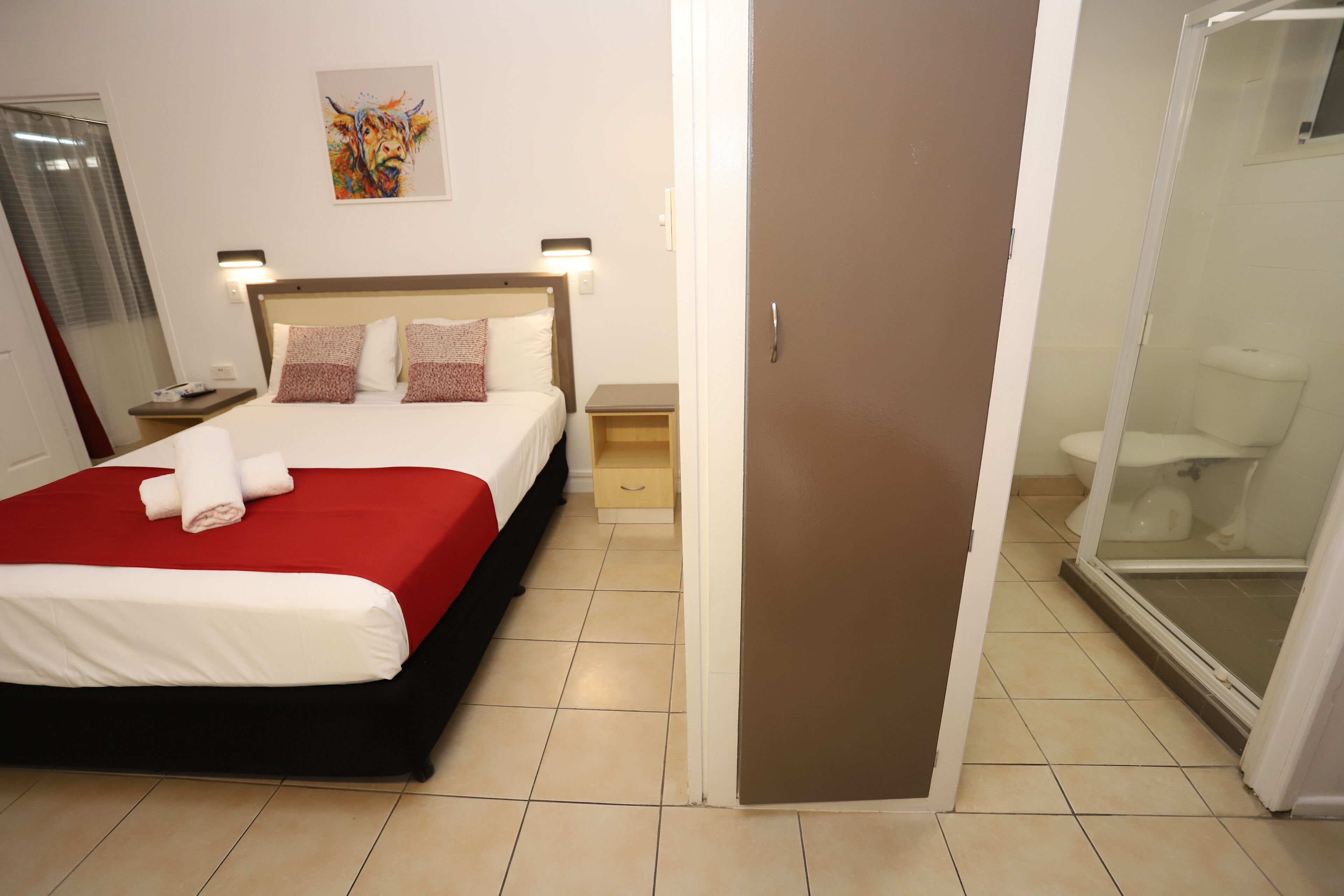 A hotel room with a bed, a nightstand, a wall-mounted lamp, a bathroom with a toilet and shower, and a door.