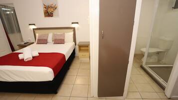 A hotel room with a bed, a nightstand, a wall-mounted lamp, a bathroom with a toilet and shower, and a door.