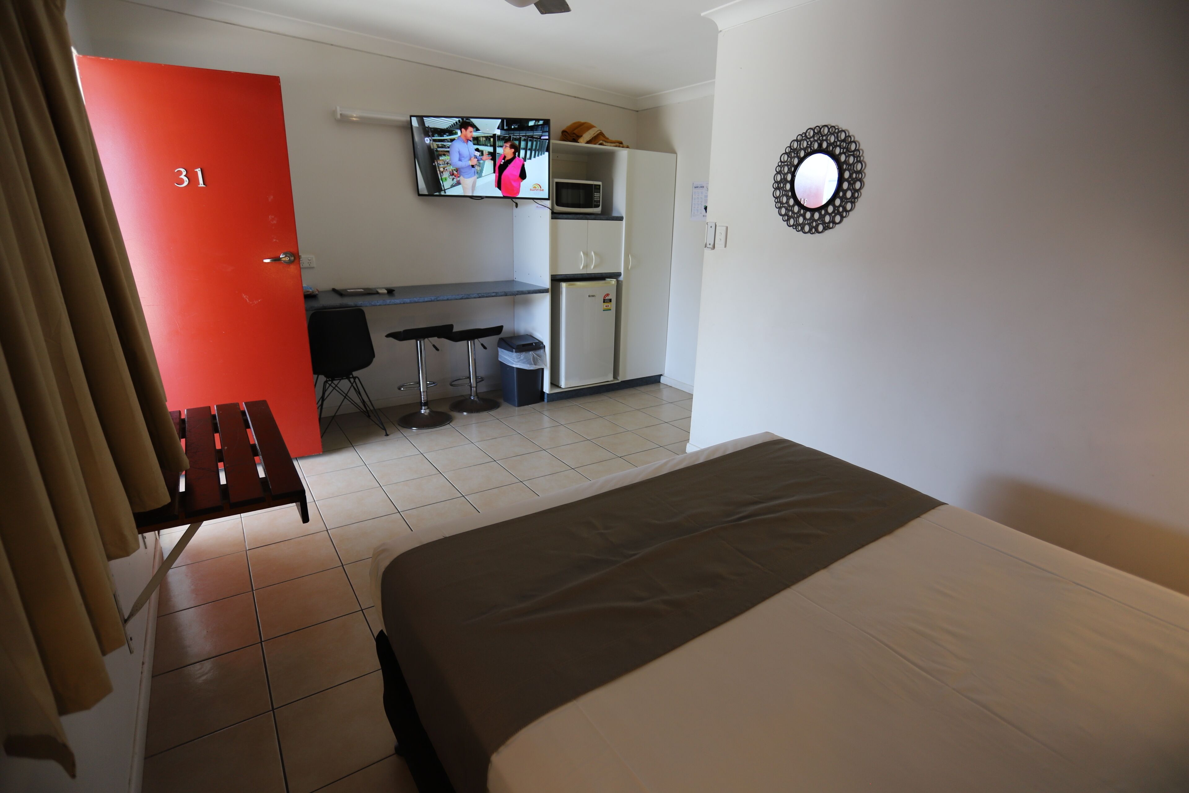 A hotel room with a bed, a desk, a chair, a trash bin, a kitchenette, and a TV on the wall.