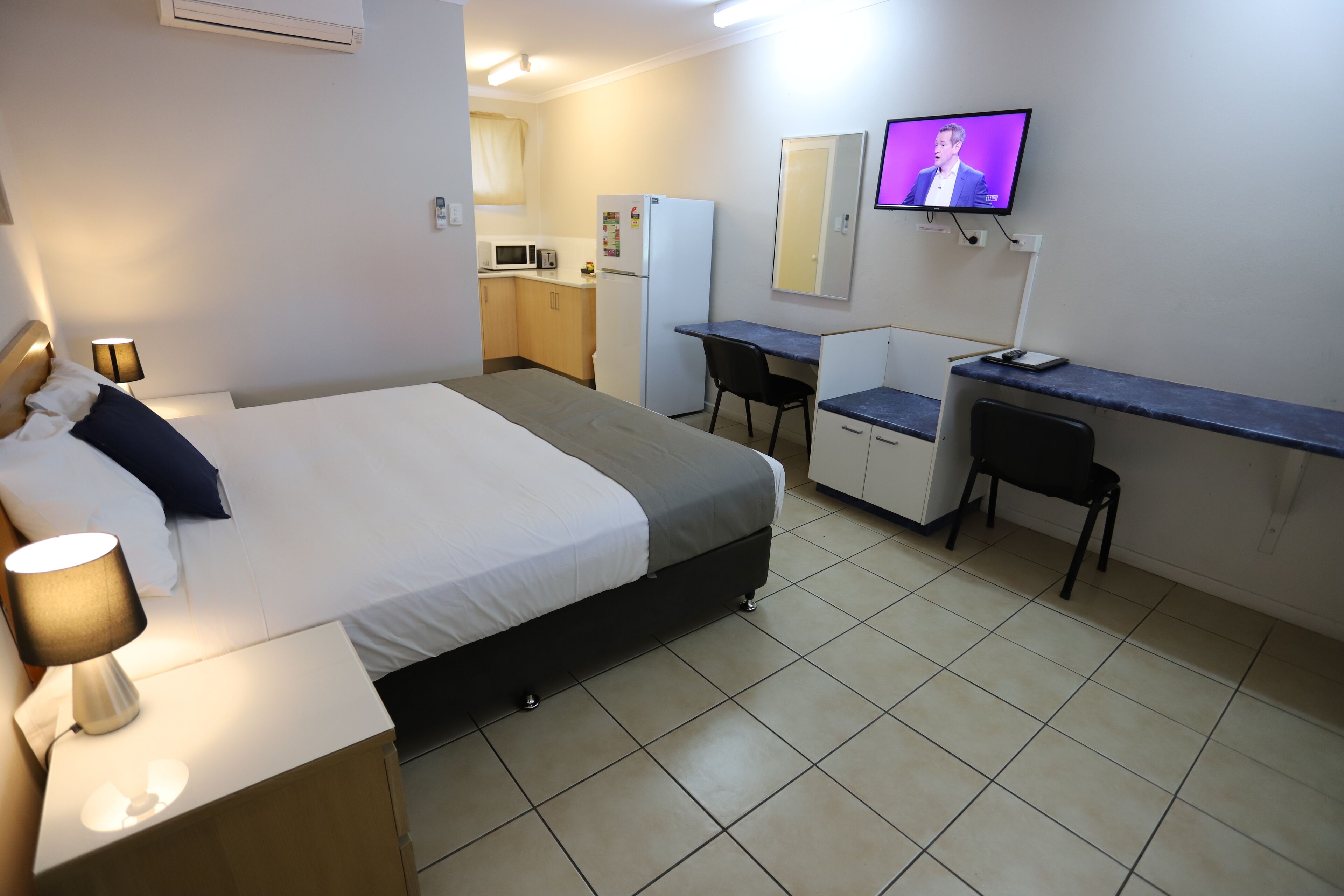 Camera d'albergo con un letto, comodino, lampada, scrivania, sedia, angolo cottura e televisore appeso al muro.