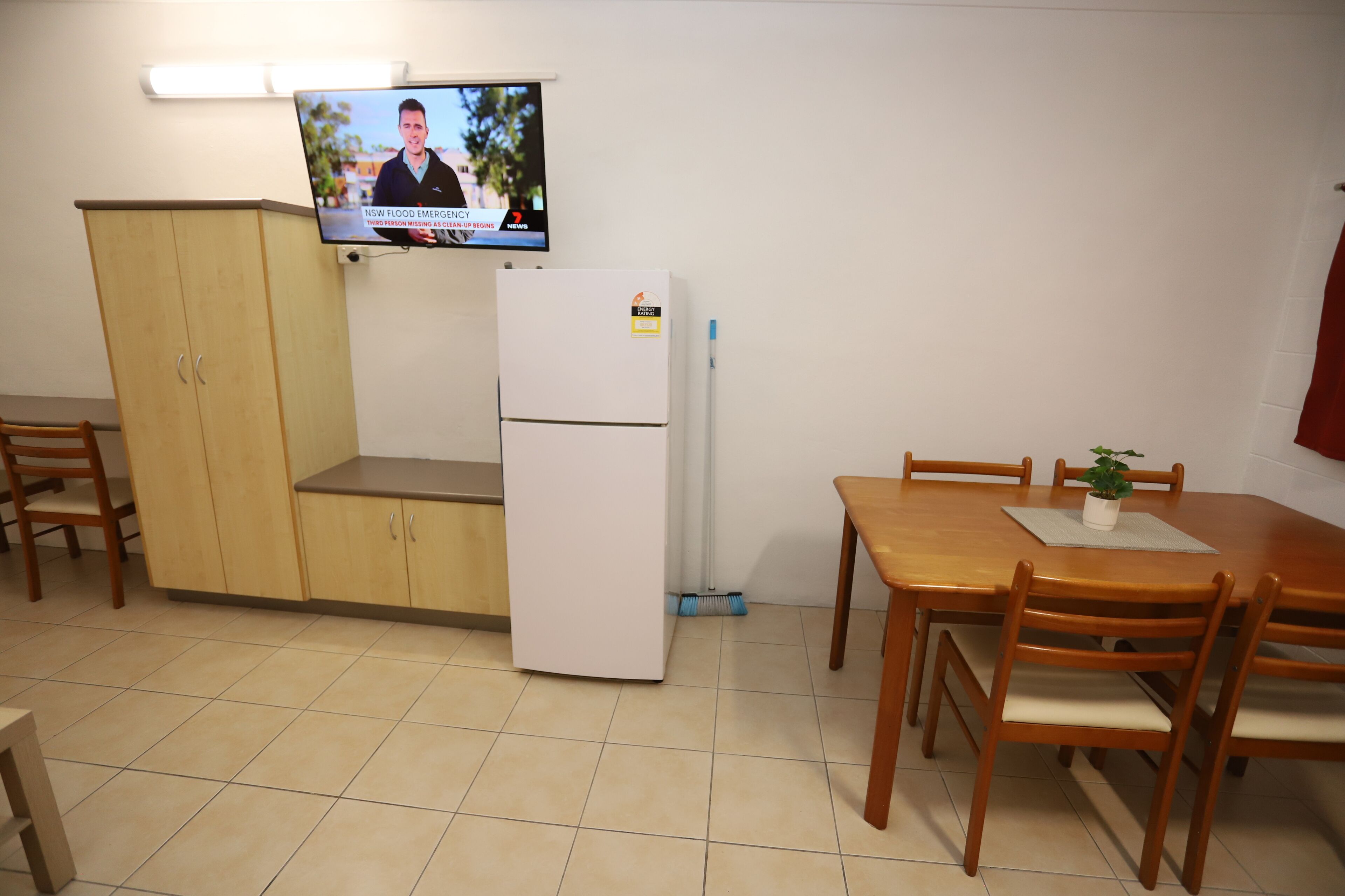 A small kitchen with a refrigerator, a wooden cabinet, a dining table with chairs, and a TV mounted on the wall displaying a news program.