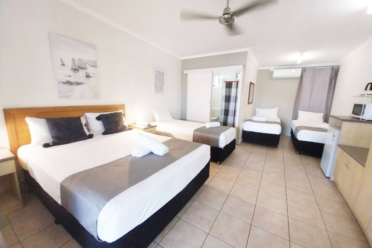 Une chambre d’hôtel avec un grand lit, deux petits lits, un bureau avec un ordinateur et un ventilateur de plafond.