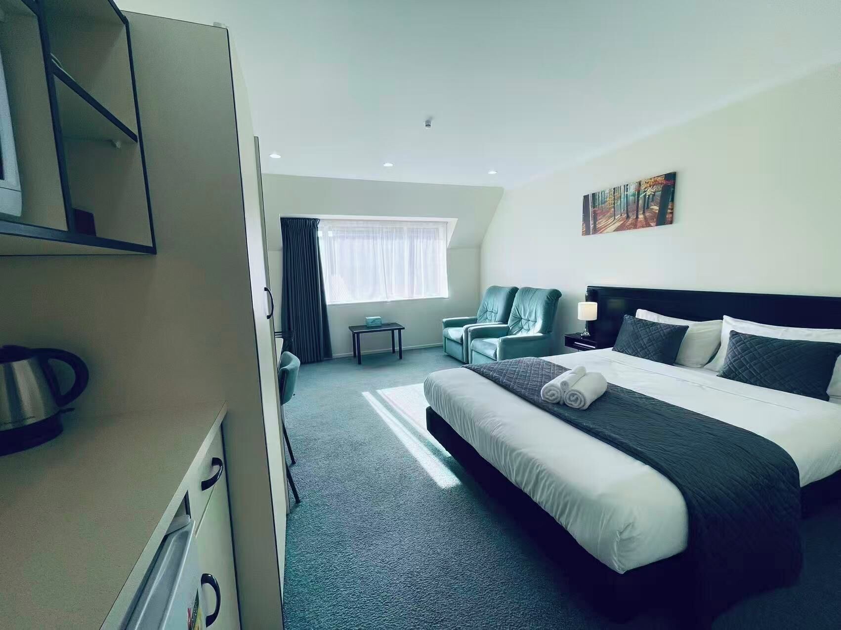 Une chambre d’hôtel avec un lit, deux fauteuils, une petite table, un bureau, une fenêtre avec des rideaux et un tableau au mur.