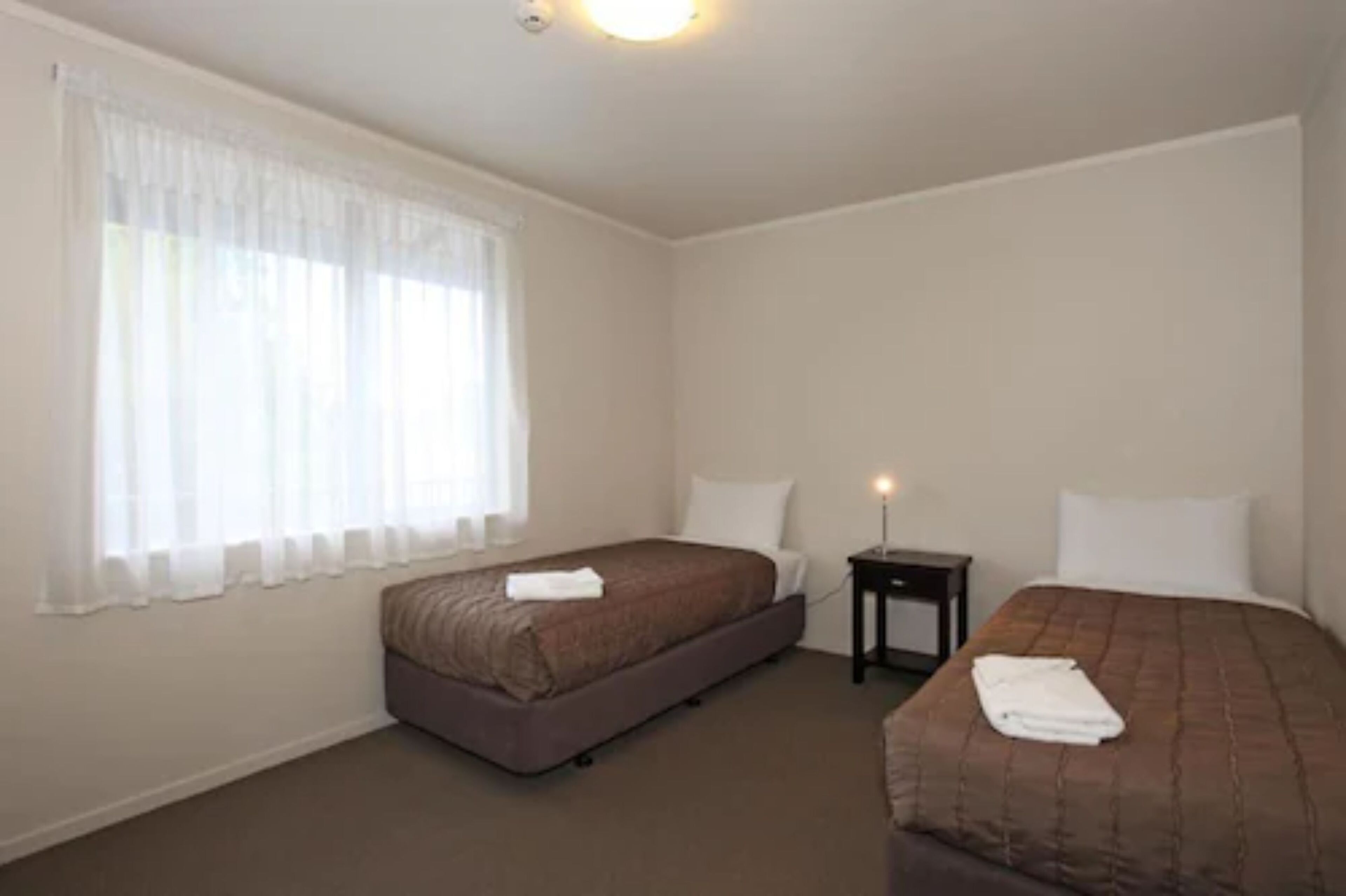 A hotel room with two beds, a small bedside table, and a window with sheer curtains.
