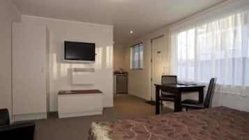 Ett hotellrum med en säng, ett skrivbord med en stol, en tv som är monterad på väggen och ett fönster med tunna, genomskinliga gardiner.
