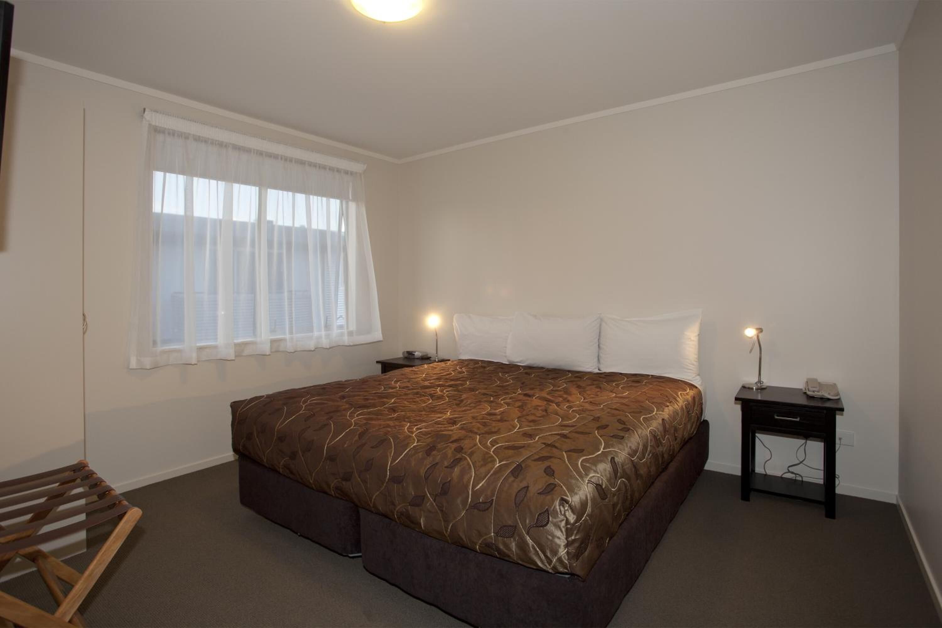 Un dormitorio con una cama grande, una mesita de noche con lámpara, una silla y una ventana con cortinas de gasa.