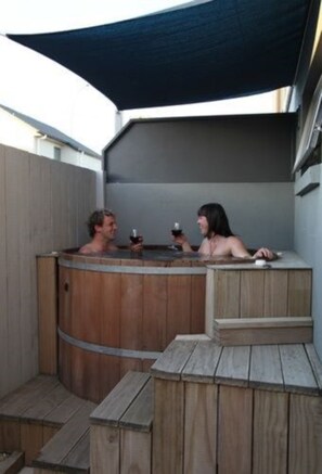 A wooden hot tub with two people inside, surrounded by a wooden deck and a large dark canopy. - Atrium on Ulster (Hamilton)