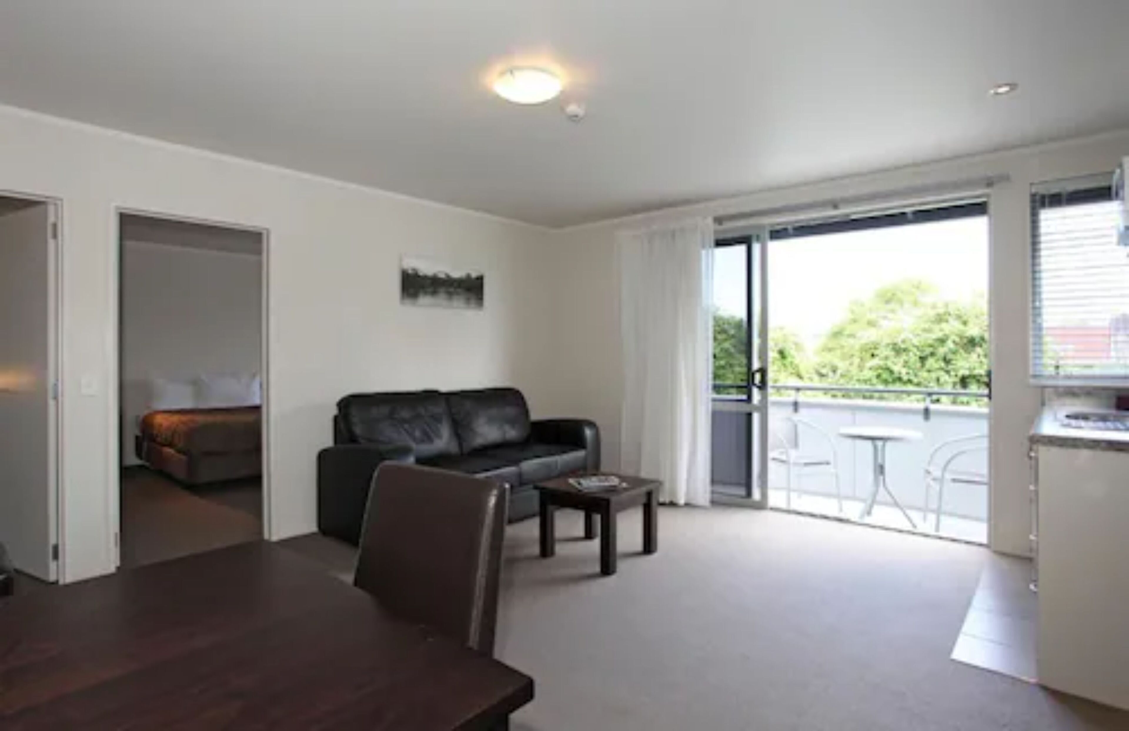 A modern living room with a dark brown dining table, a black leather sofa, and a small coffee table. There is a sliding glass door leading to a balcony with outdoor furniture.