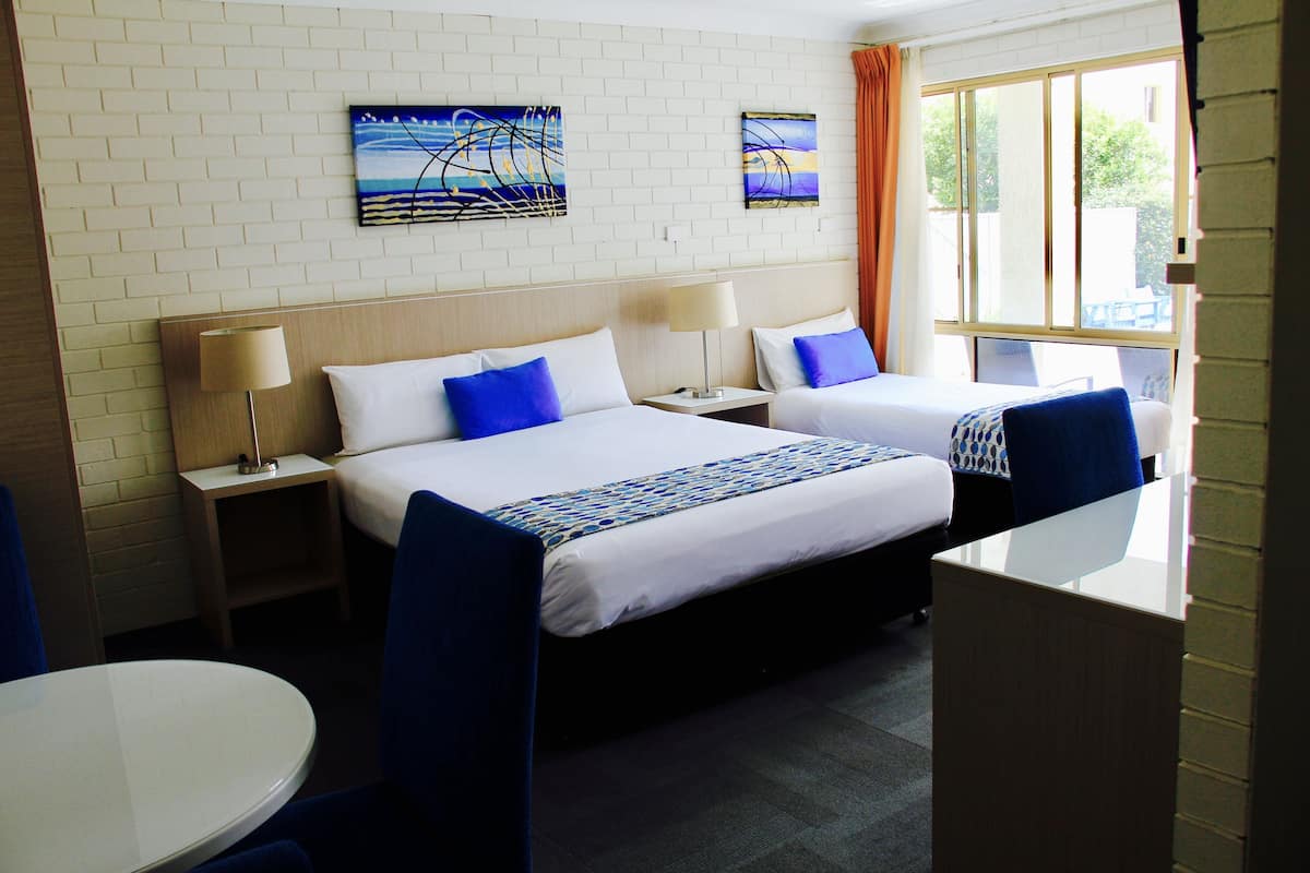 A hotel room with two beds, a desk, and a chair. There are two paintings on the wall and a window with curtains.