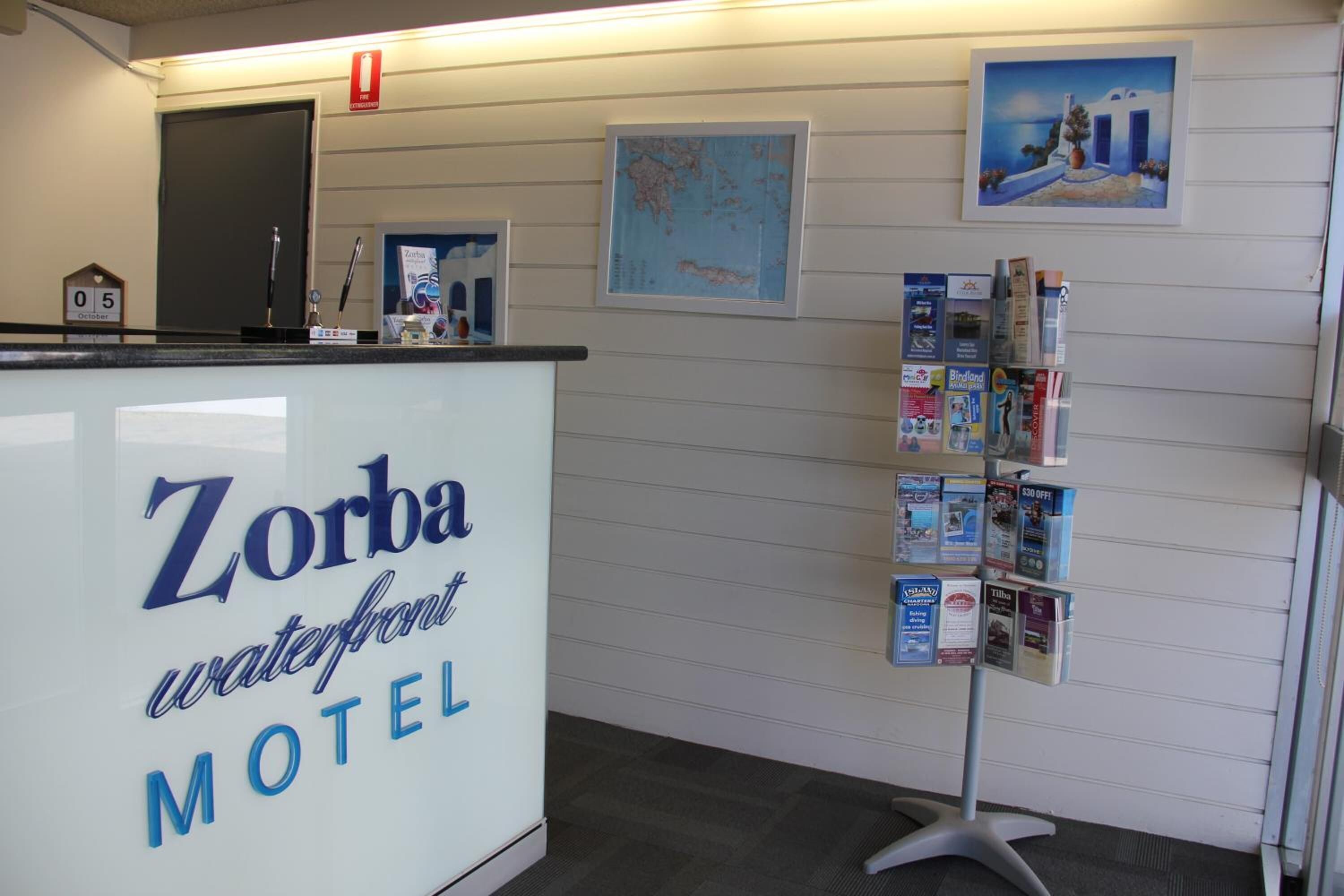 Une réception avec une enseigne indiquant « Zorba Waterfront Motel », une carte affichée au mur et un présentoir rempli de brochures.