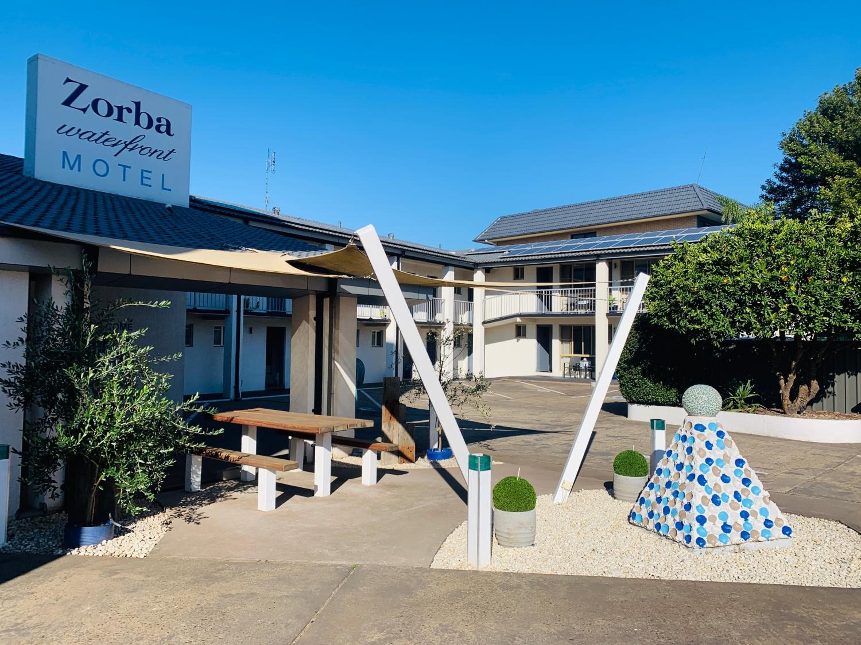 The exterior of the Zorba Waterfront Motel with a clear sky, a signboard, a patio area with benches, and a decorative sculpture.