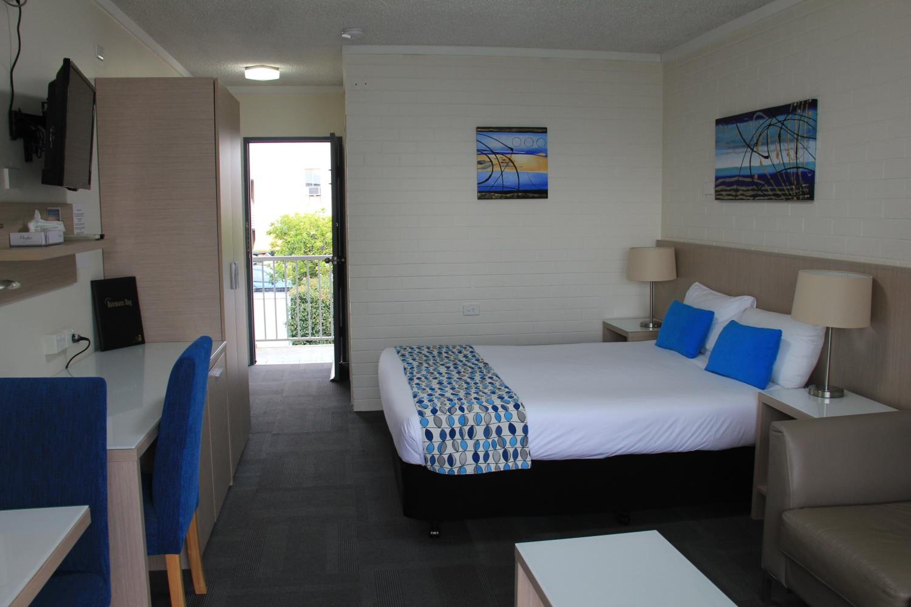Ein Hotelzimmer mit einem Bett, Nachttischen, einem Schreibtisch und Stühlen. An der Wand hängen zwei Gemälde.