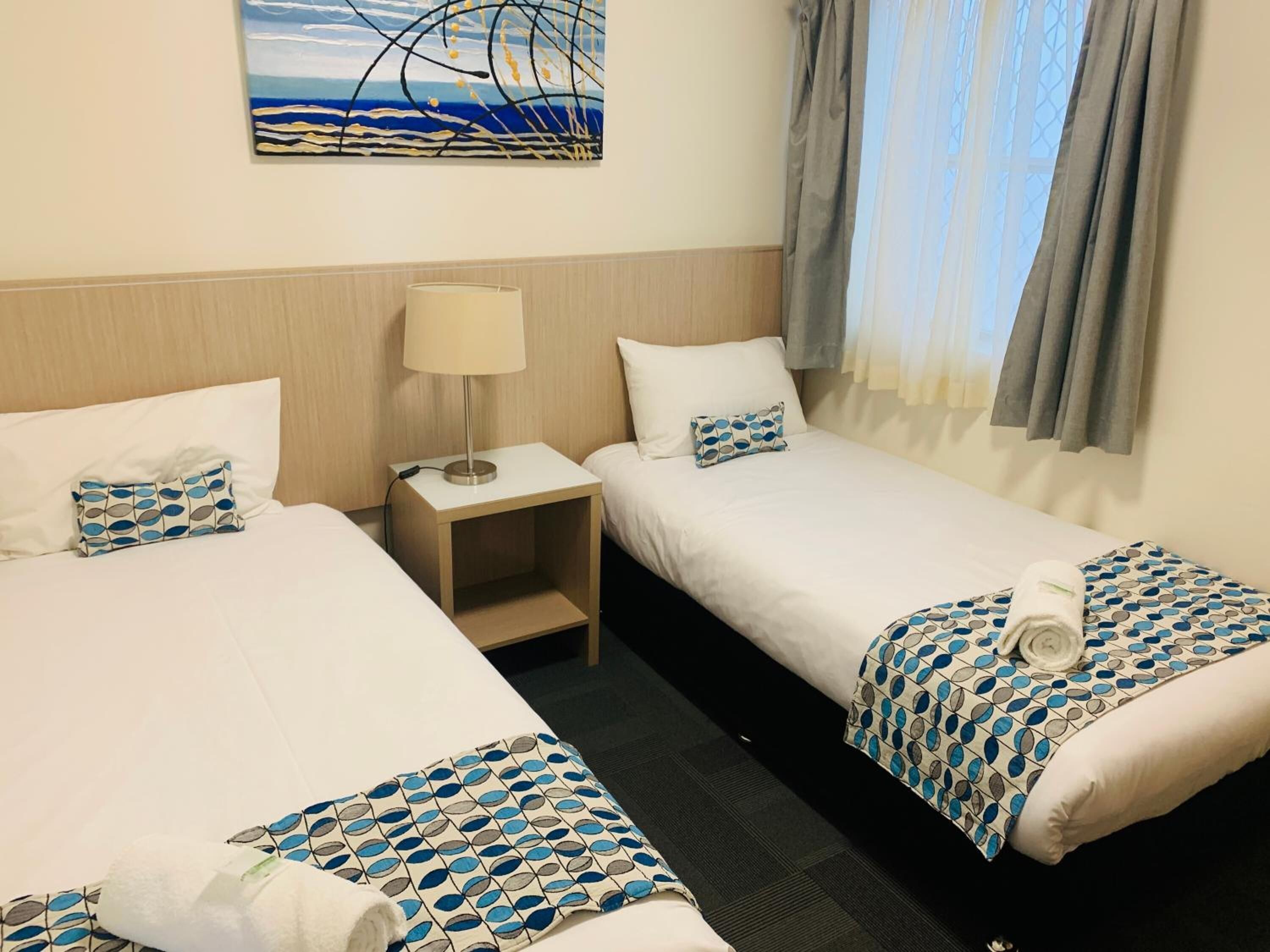 A hotel room with two single beds, a nightstand with a lamp, a painting on the wall, and a window with curtains.