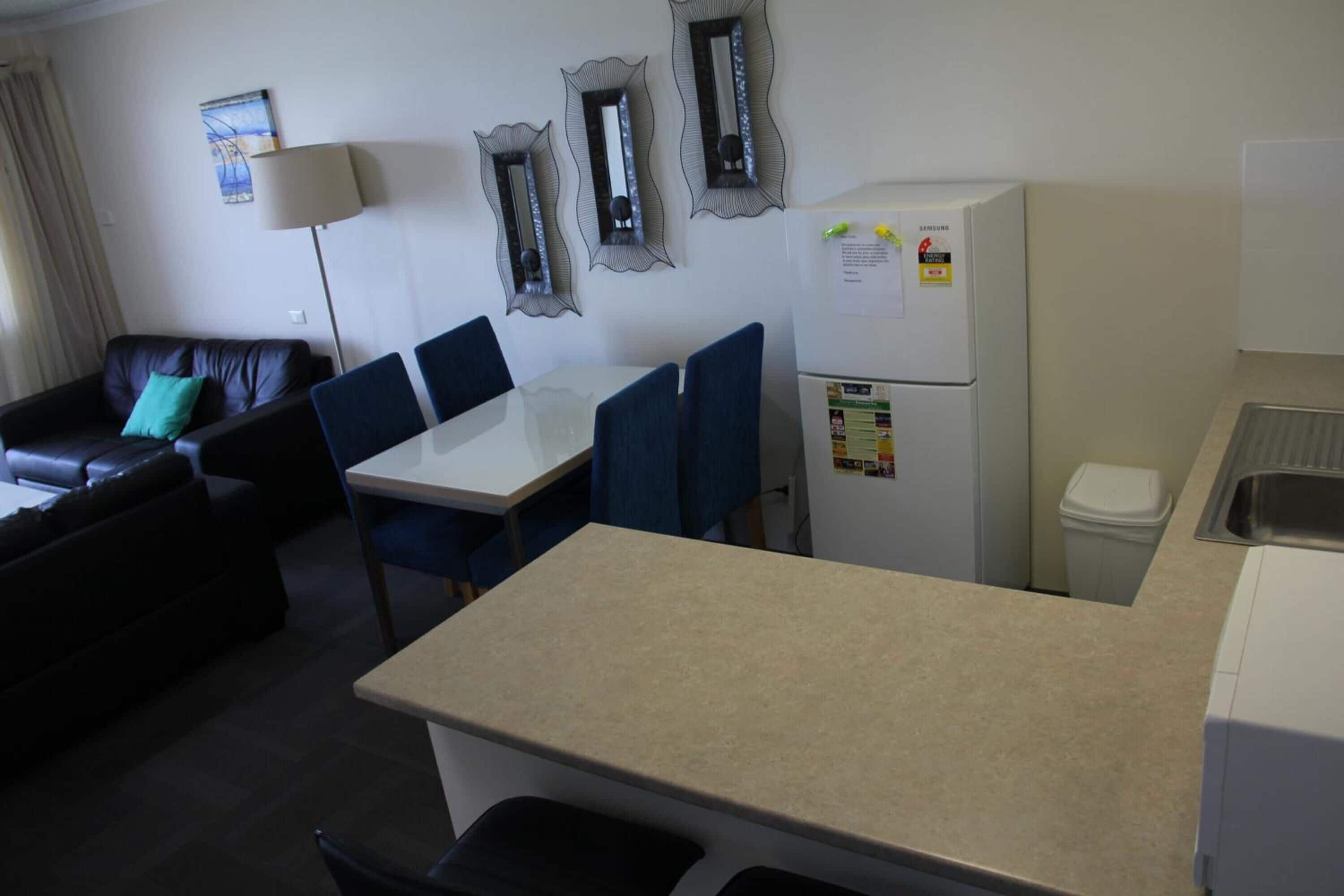 Habitación de hotel compacta con kitchenette, una zona de comedor con mesa y sillas, un sofá, una lámpara y espejos decorativos en la pared.