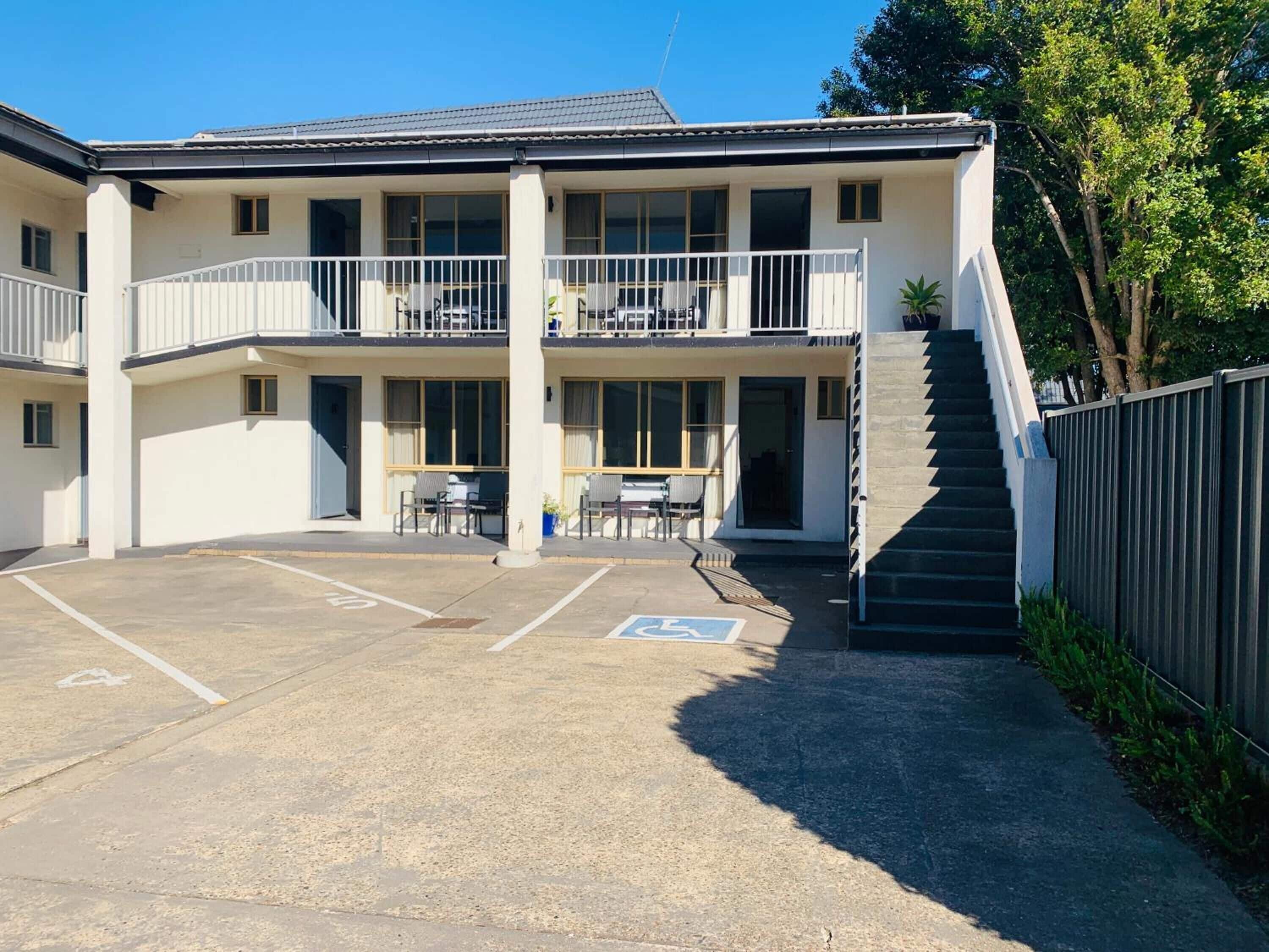 Un bâtiment à deux étages doté d’un balcon à l’étage supérieur, d’un espace extérieur couvert avec des sièges et d’un parking comprenant une place réservée aux personnes handicapées.