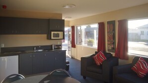 A compact kitchen with dark cabinets, a microwave, and a sink. There is a dining area with a table and two chairs, and a window with red curtains. - Phoenix Motel (Temuka)