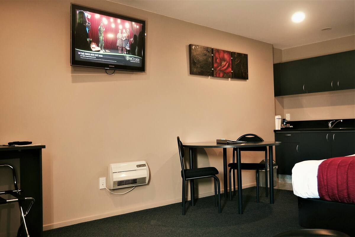 A hotel room with a TV mounted on the wall, a dining table with chairs, a bed with a red blanket, a kitchenette, and an air conditioning unit.