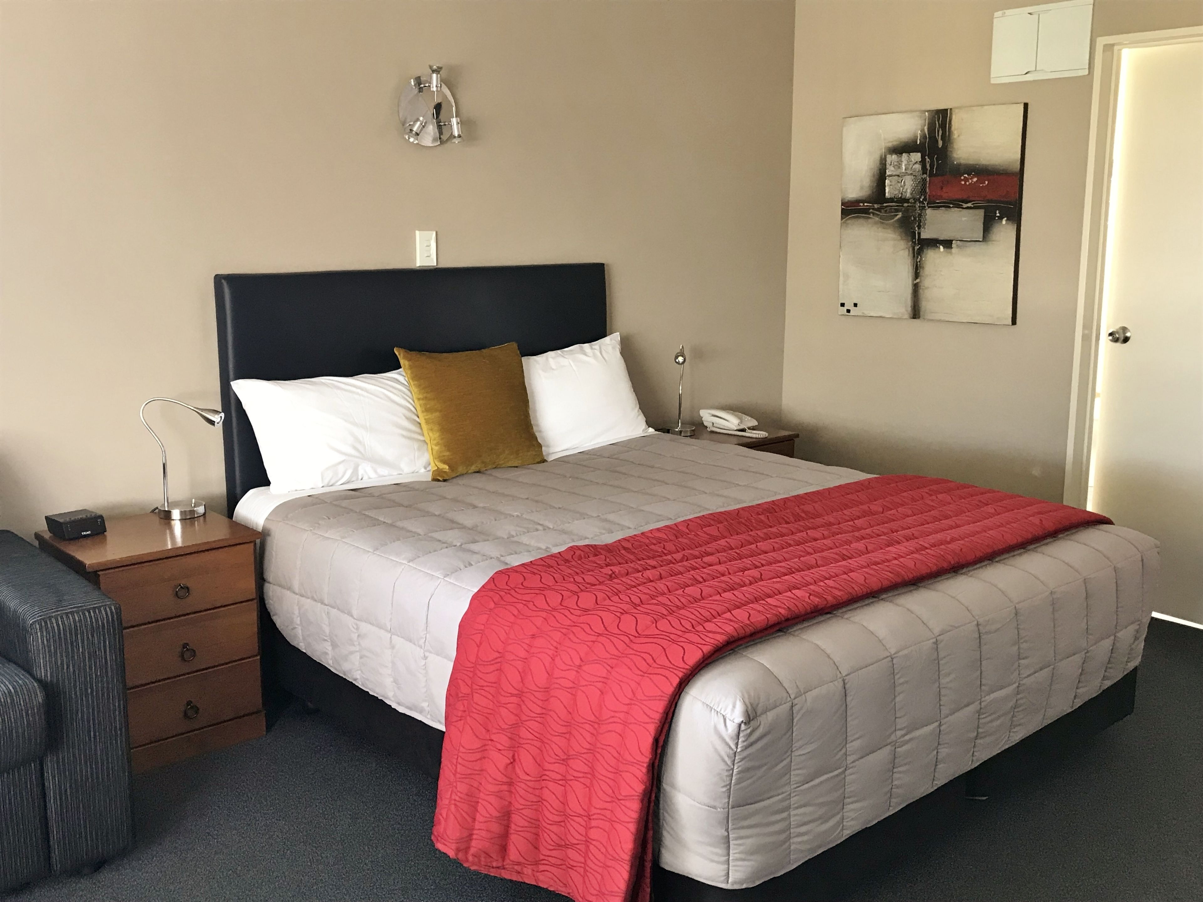 A neatly made bed with a red blanket, a bedside lamp, and a nightstand with a phone and a box.