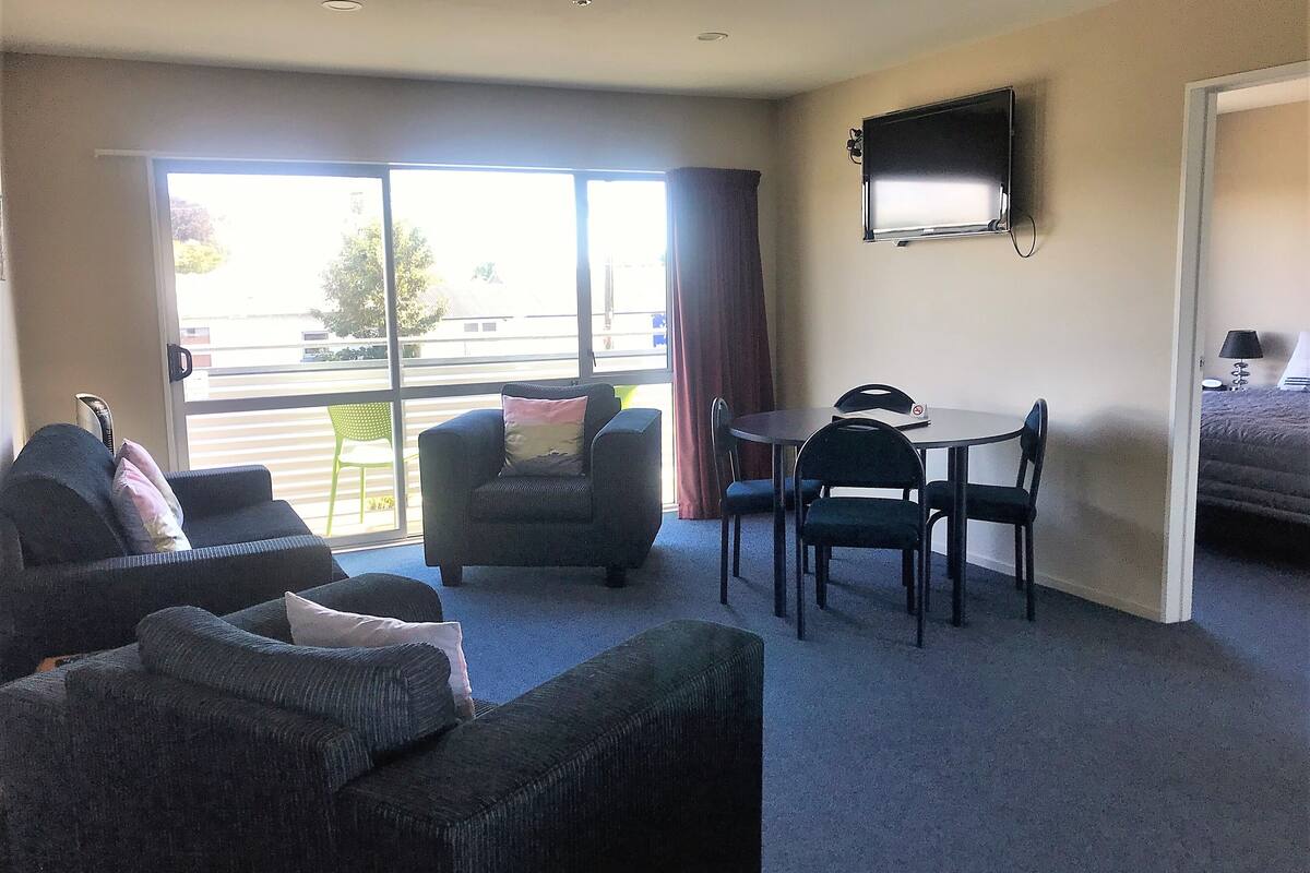 A hotel room with a sofa, armchair, and a small dining table with chairs. There is a TV mounted on the wall and a window with curtains.