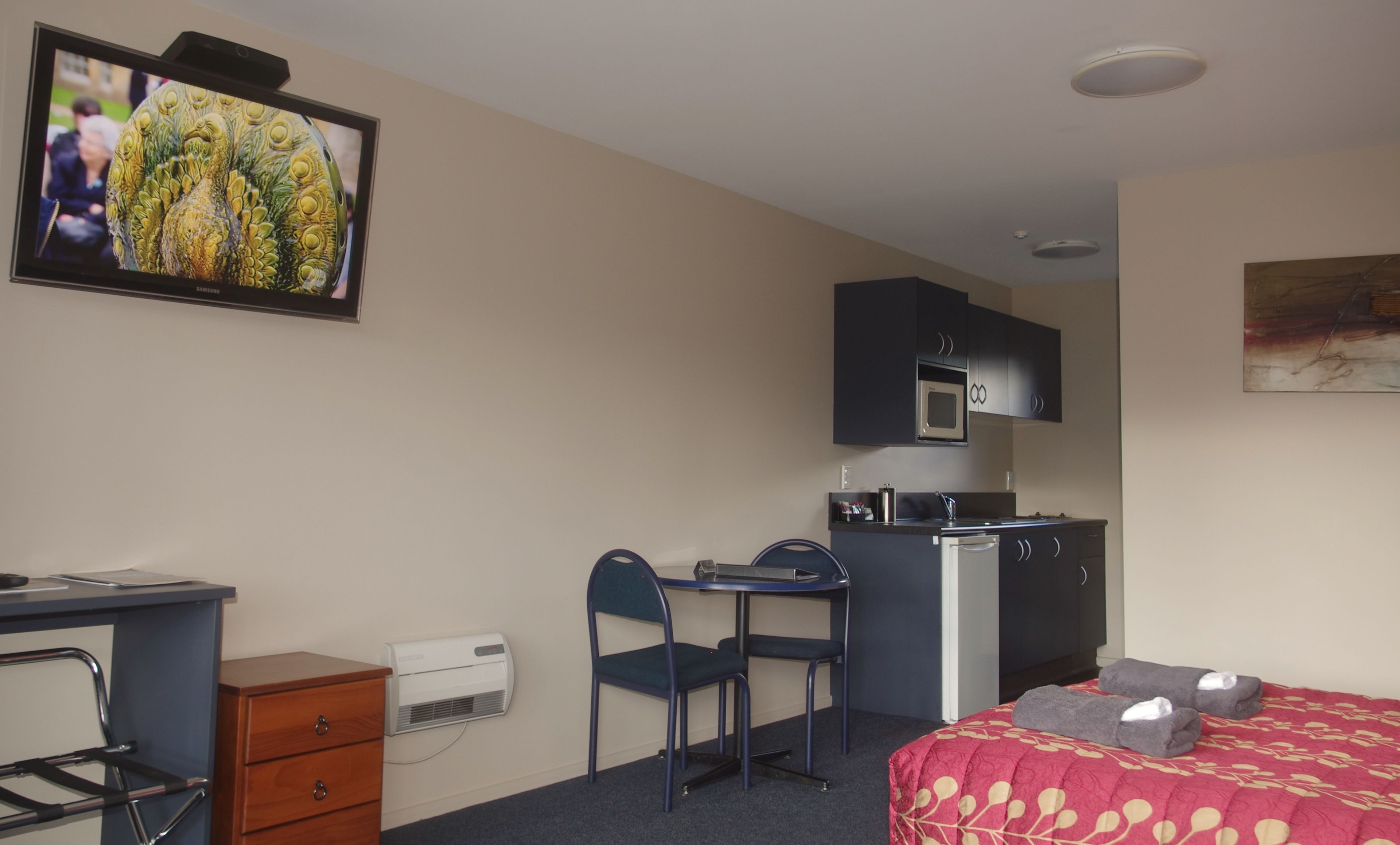 A compact hotel room with a bed, desk, chair, kitchenette, and a TV mounted on the wall.