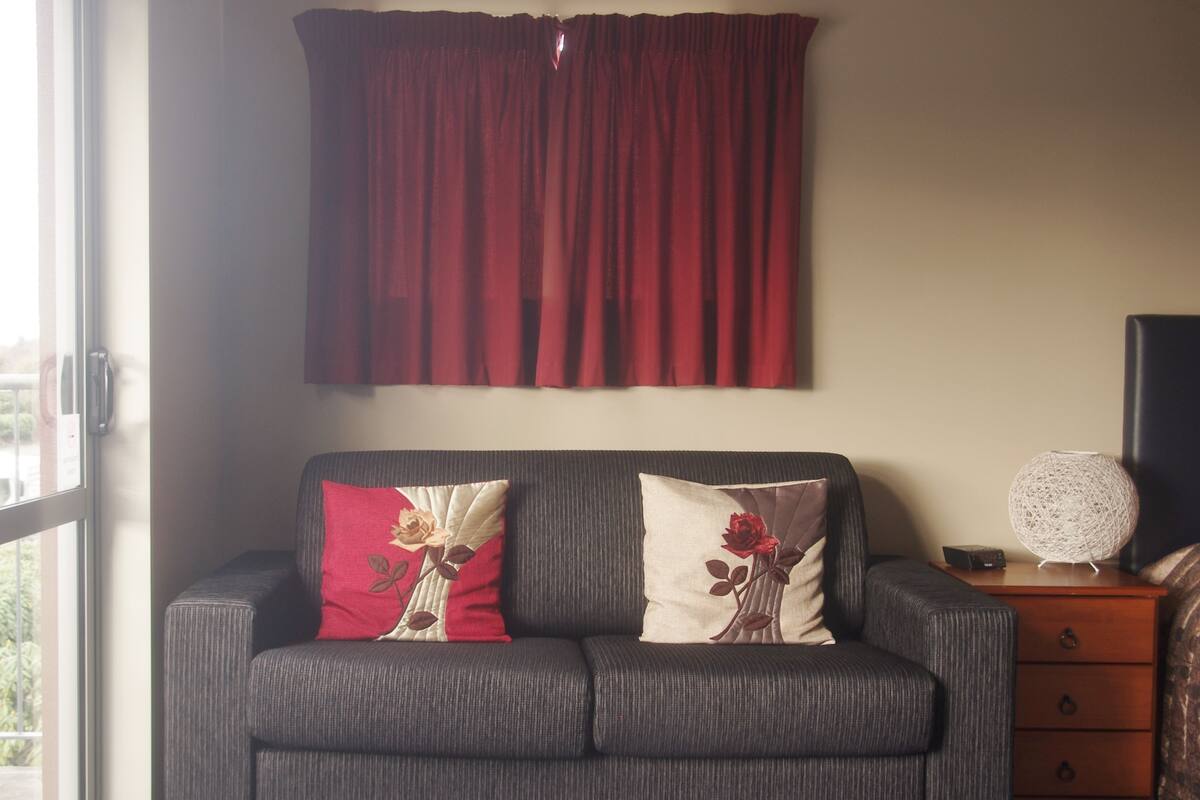 A grey sofa with two decorative pillows, a red curtain, a small table with a spherical lamp, and a television on a stand.