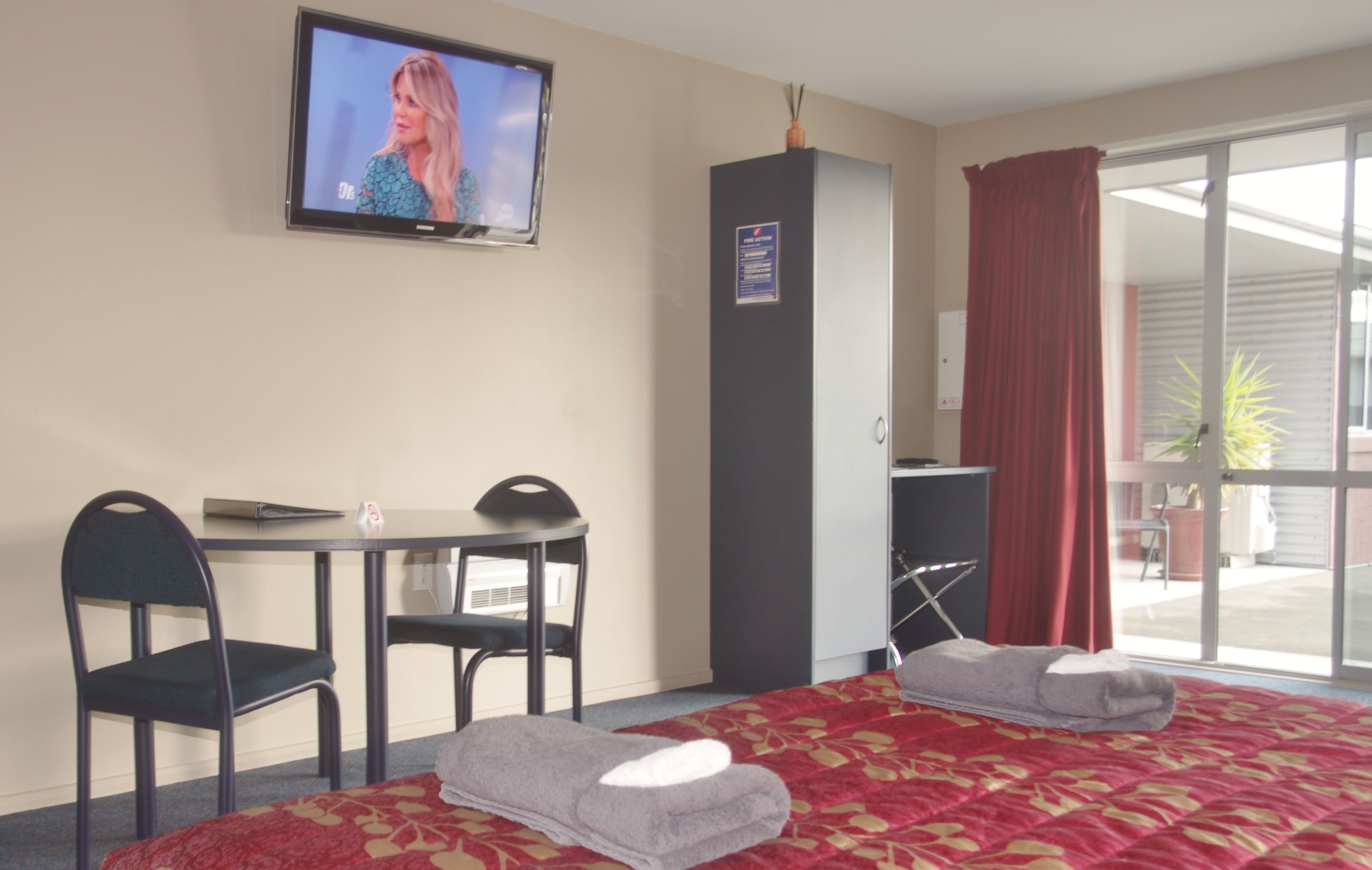 Habitación de hotel con cama, dos sillas, una mesita, televisor, armario y puerta corrediza de vidrio al exterior.