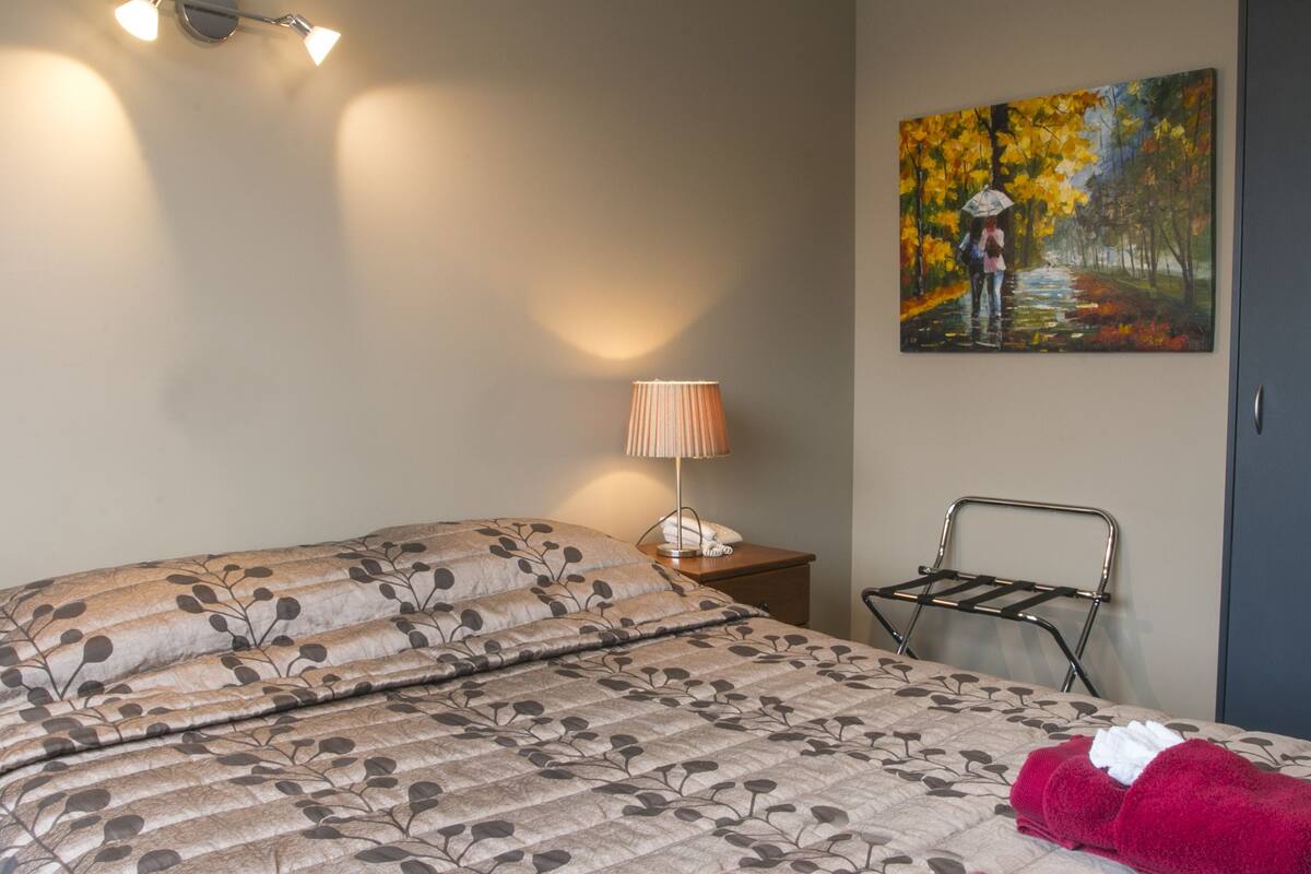A neatly made bed with a floral-patterned bedspread, a bedside lamp, and a painting of a couple walking in a park.