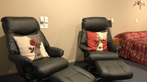 Two black leather recliner chairs with footrests, a beige wall with a power outlet, and a red floral bedspread. - Phoenix Motel (Temuka)