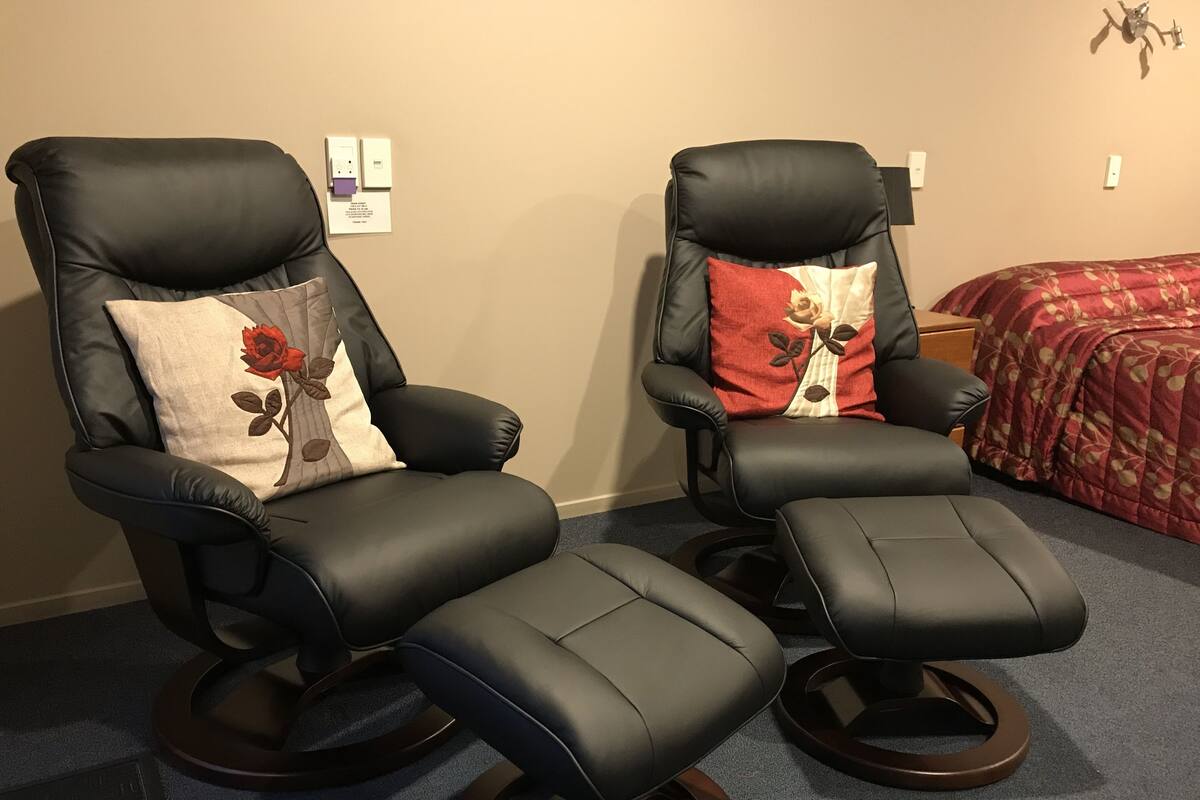 Two black leather recliner chairs with footrests, a beige wall with a power outlet, and a red floral bedspread.