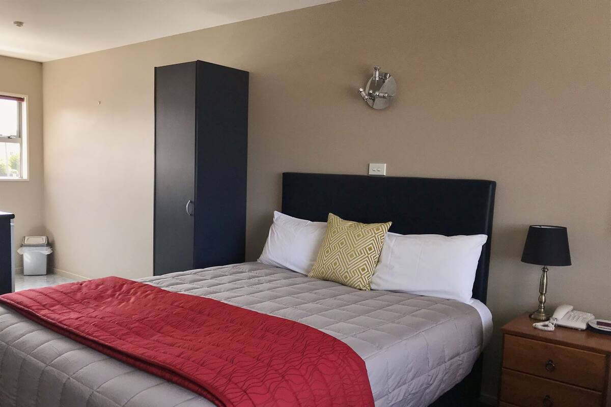 A neatly made bed with a red blanket, a bedside lamp, and a phone on a wooden nightstand.
