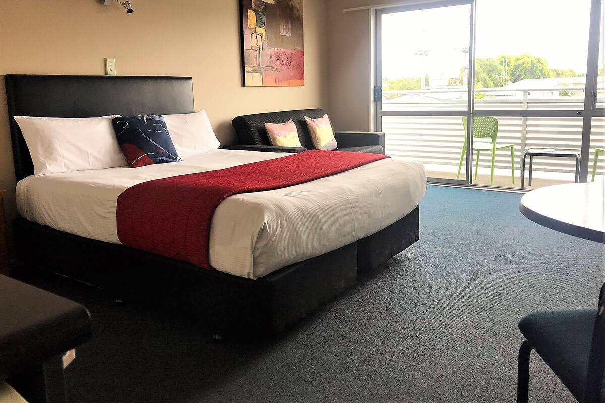 A hotel room with a large bed, a red throw blanket, two armchairs, a small table, and a large window with a view of the outside.