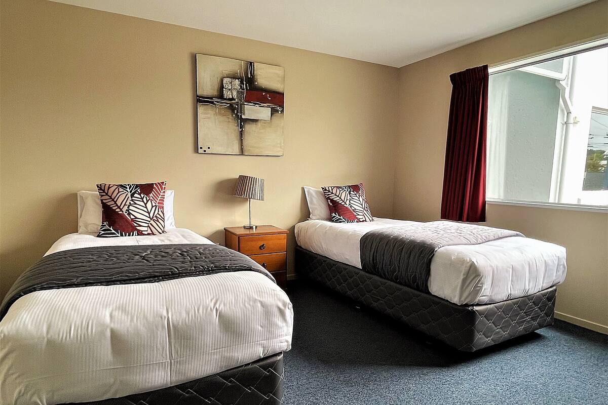 A hotel room with two beds, a nightstand, a lamp, and a window with curtains.