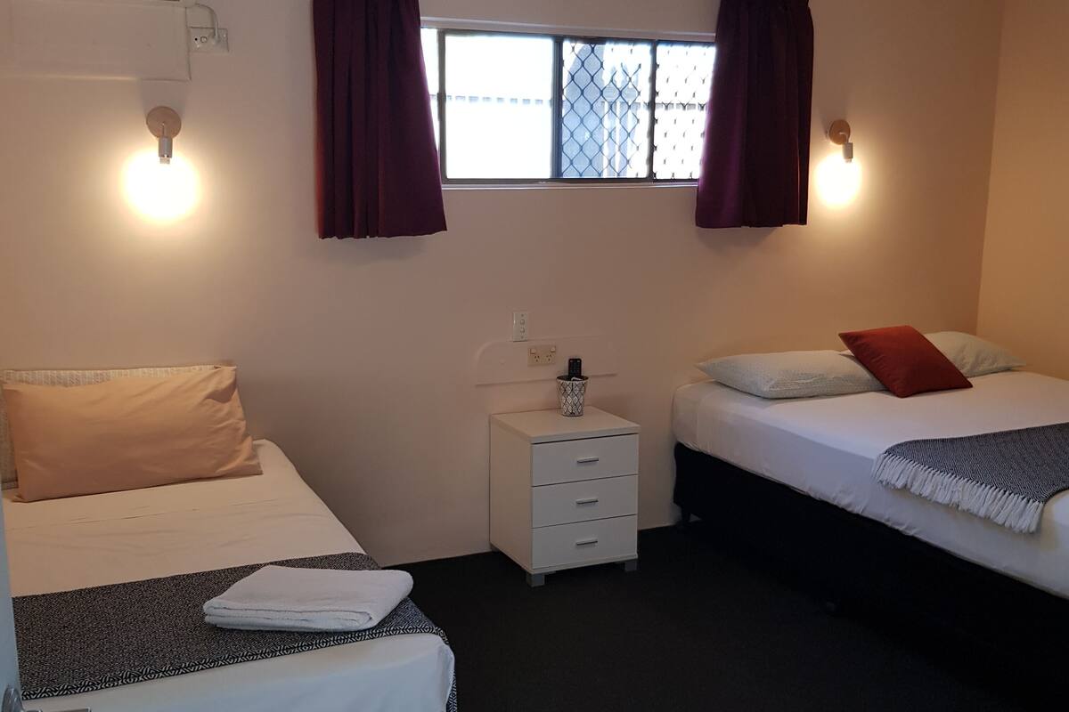 A small, clean room with two beds, a nightstand, a window with curtains, and a wall-mounted light fixture.