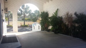 Una entrada con pared blanca y un arco, que conduce a un estacionamiento con palmeras y un pequeño edificio.
