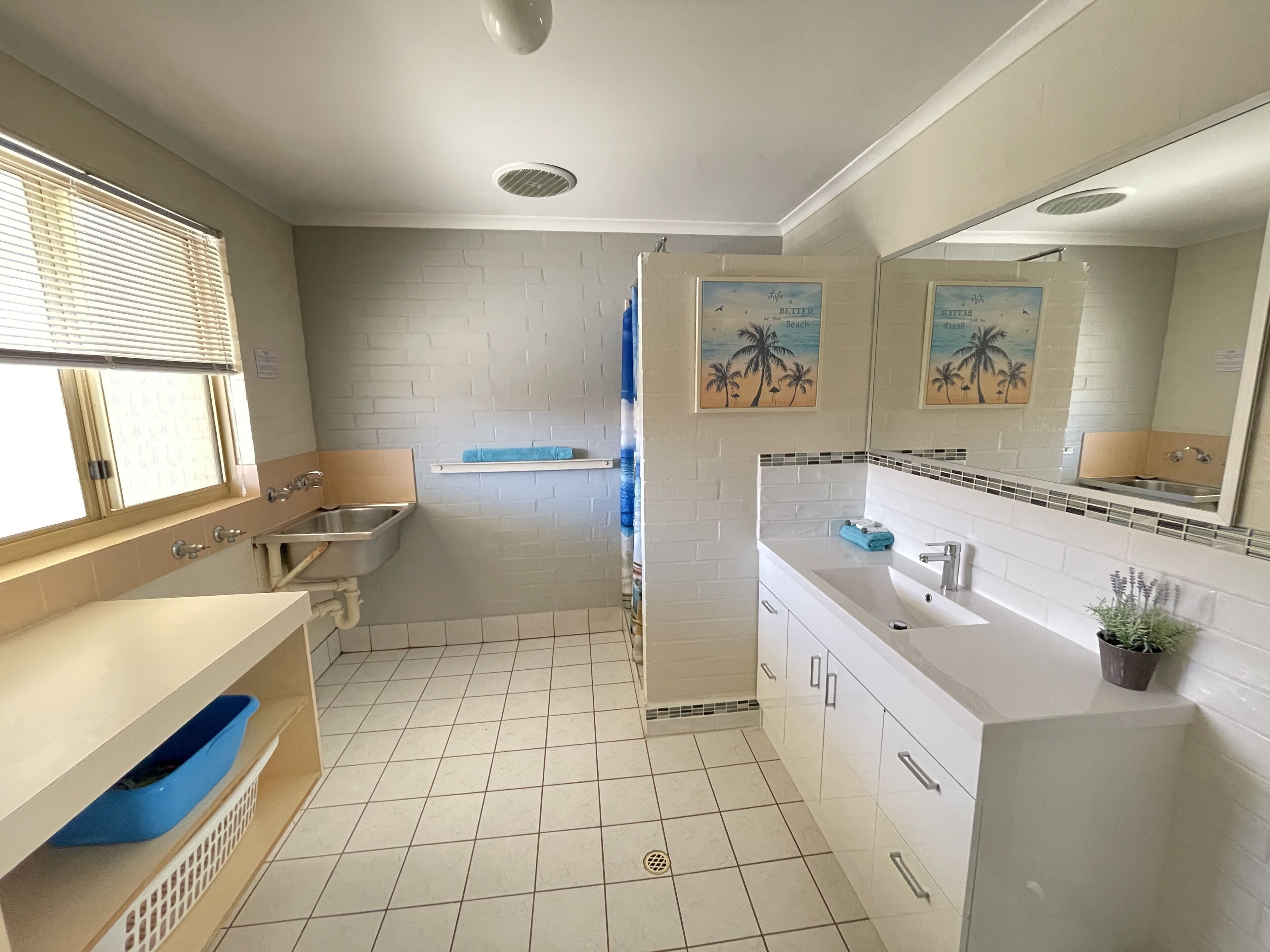 A modern bathroom with a white tiled floor, a sink with a blue towel, a mirror, and a window with blinds.