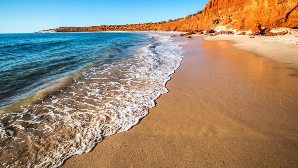 Una playa con agua azul cristalina, orilla de arena y un acantilado rocoso.