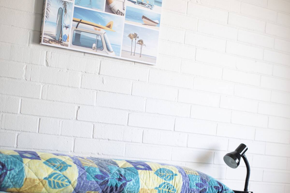 Un dormitorio con pared de ladrillo blanco, cama con colcha colorida, un cuadro colgado en la pared y una lámpara de escritorio.