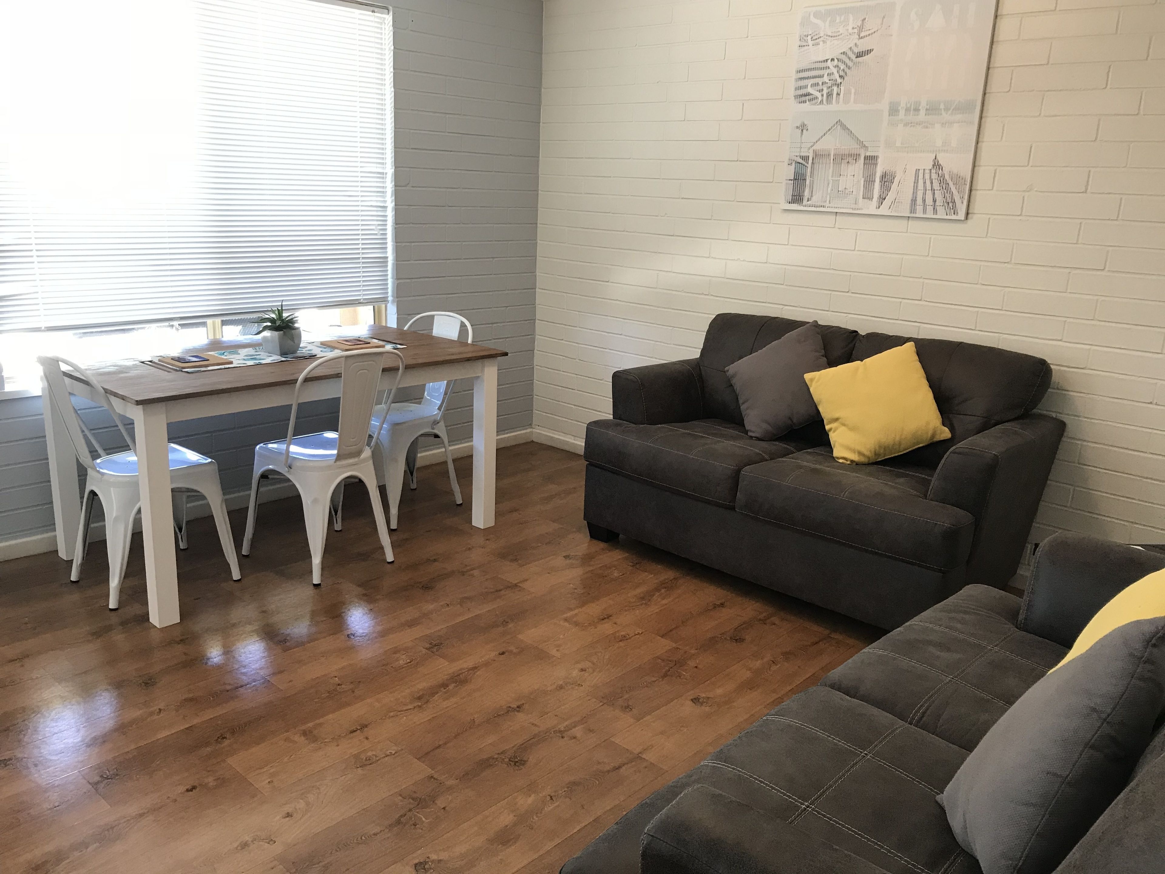 Een woonkamer met een houten eettafel, witte stoelen, een donkergrijze bank met een geel kussen en een grijze slaapbank.