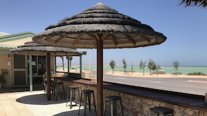 Un bar frente al mar con sombrillas de paja y paredes de piedra, con vista a una playa y una carretera.